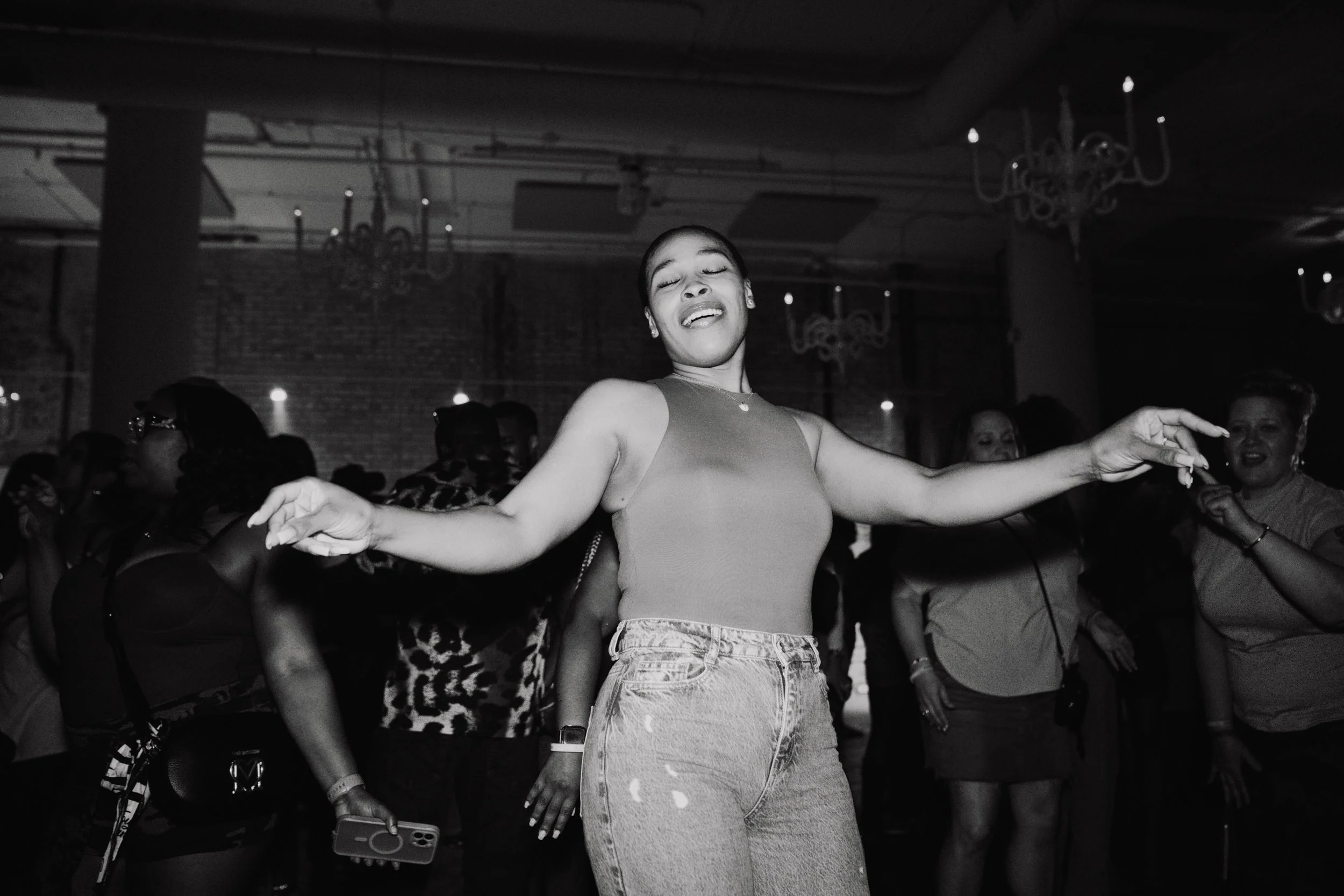 A woman dancing with her eyes closed and arms outstretched in a crowded party or dance event. The setting is indoors with chandeliers hanging from the ceiling.