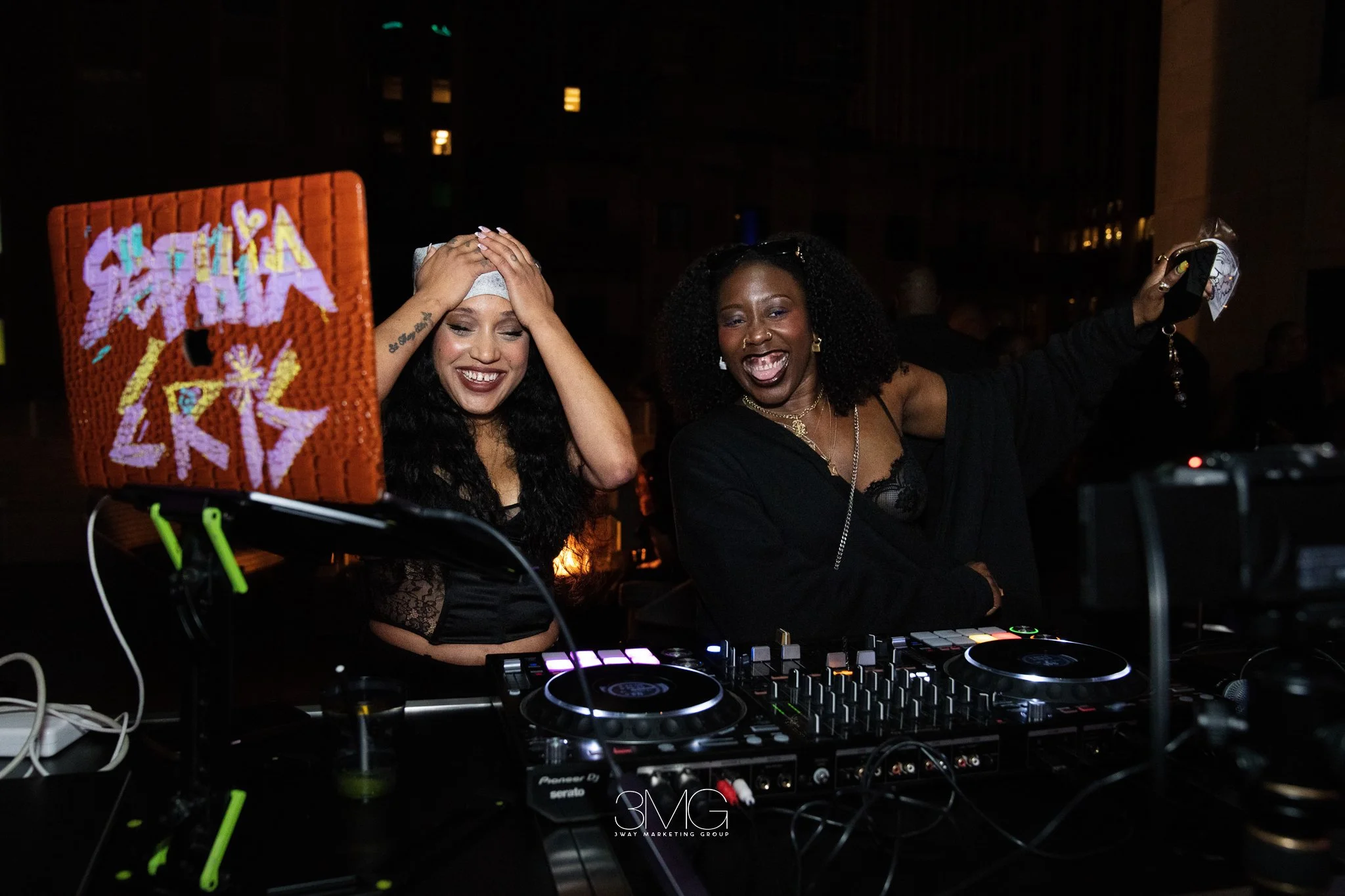 Two women at a nighttime outdoor event, one with long dark hair and a hat holding her head, the other with curly hair, wearing a black outfit, smiling and holding up a phone. There is DJ equipment in front of them, and a bright, colorful sign in the 