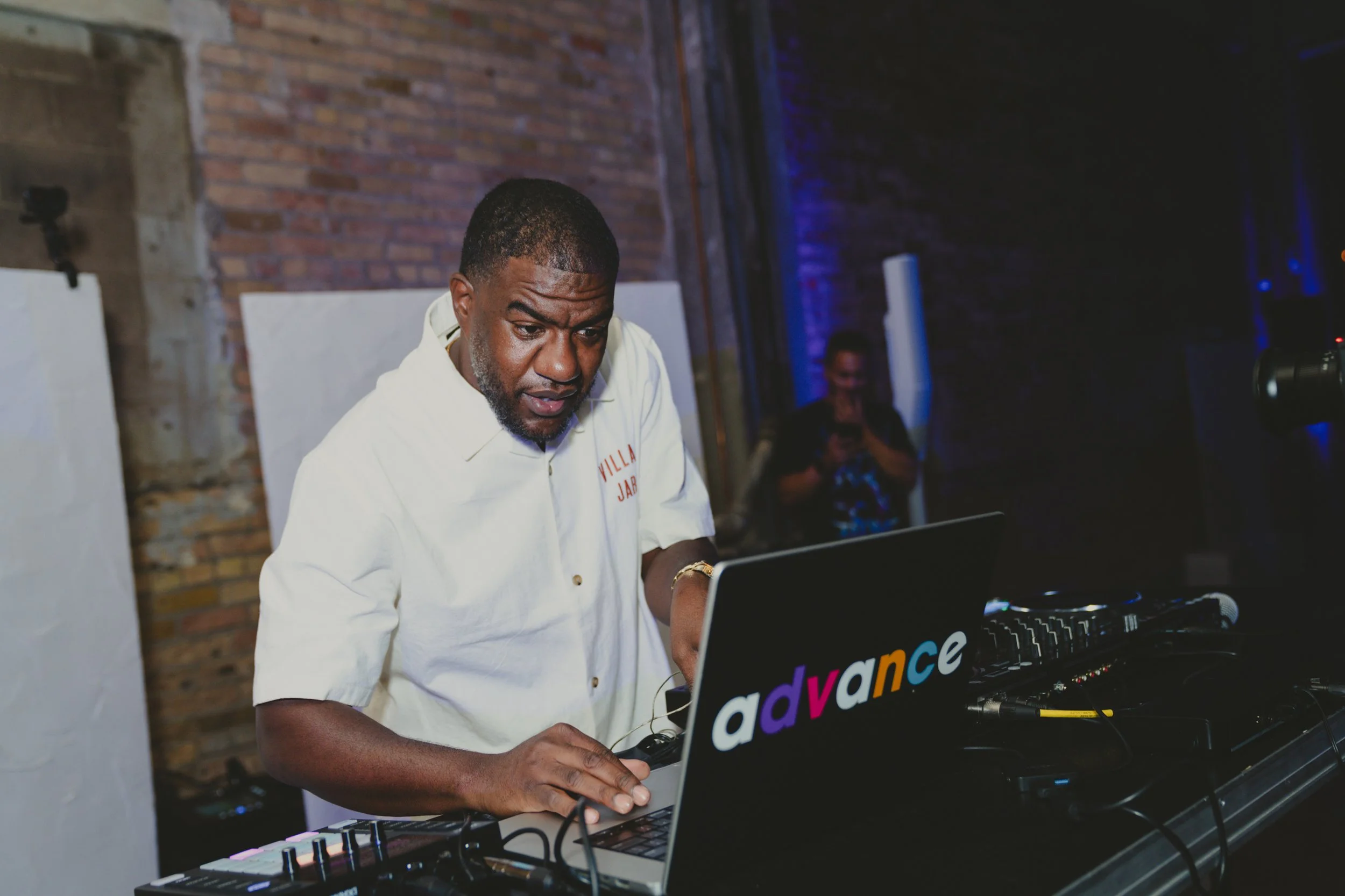 A man with short black hair and a beard, wearing a white shirt, is DJing at an event with a laptop and DJ equipment. The back wall has exposed brick, and there is a woman in the background smiling. The lighting is dim with blue accent lights.