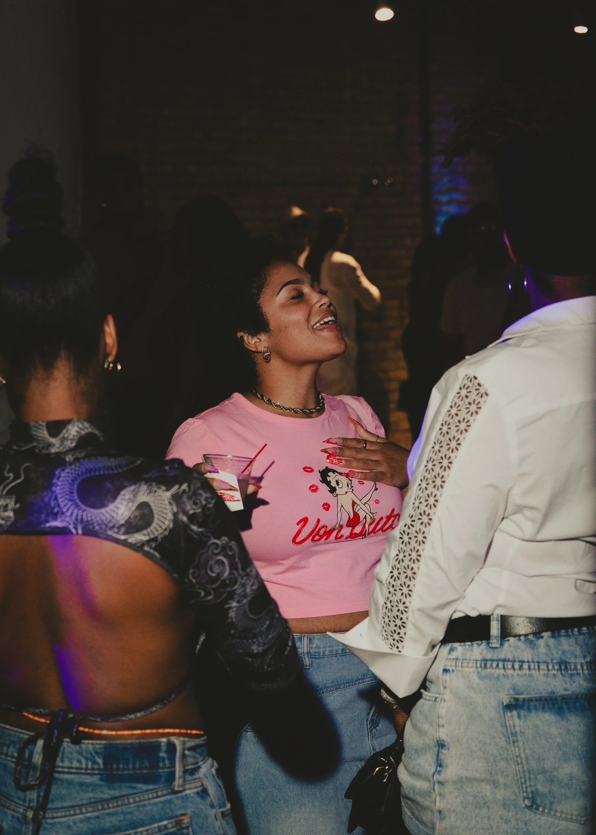 A group of people at a social gathering, mostly women, with one woman smiling and wearing a pink Betty Boop t-shirt. The setting appears to be indoors with dim lighting.