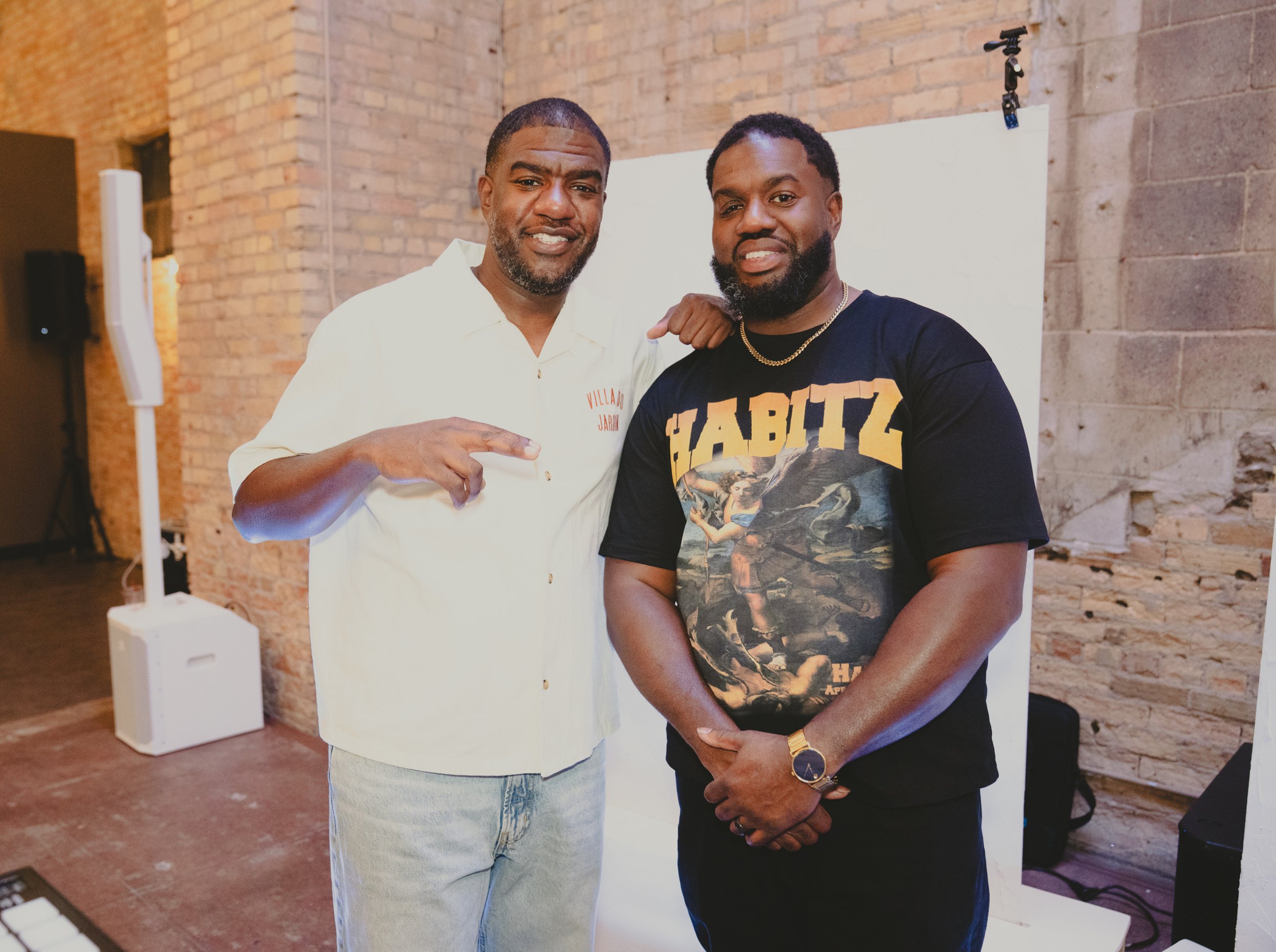 Two men standing together indoors with a brick wall background. One man is wearing a white shirt with red text and the other is wearing a black T-shirt featuring artwork, gold chain, and a watch. They are smiling and the man in black is making a gest