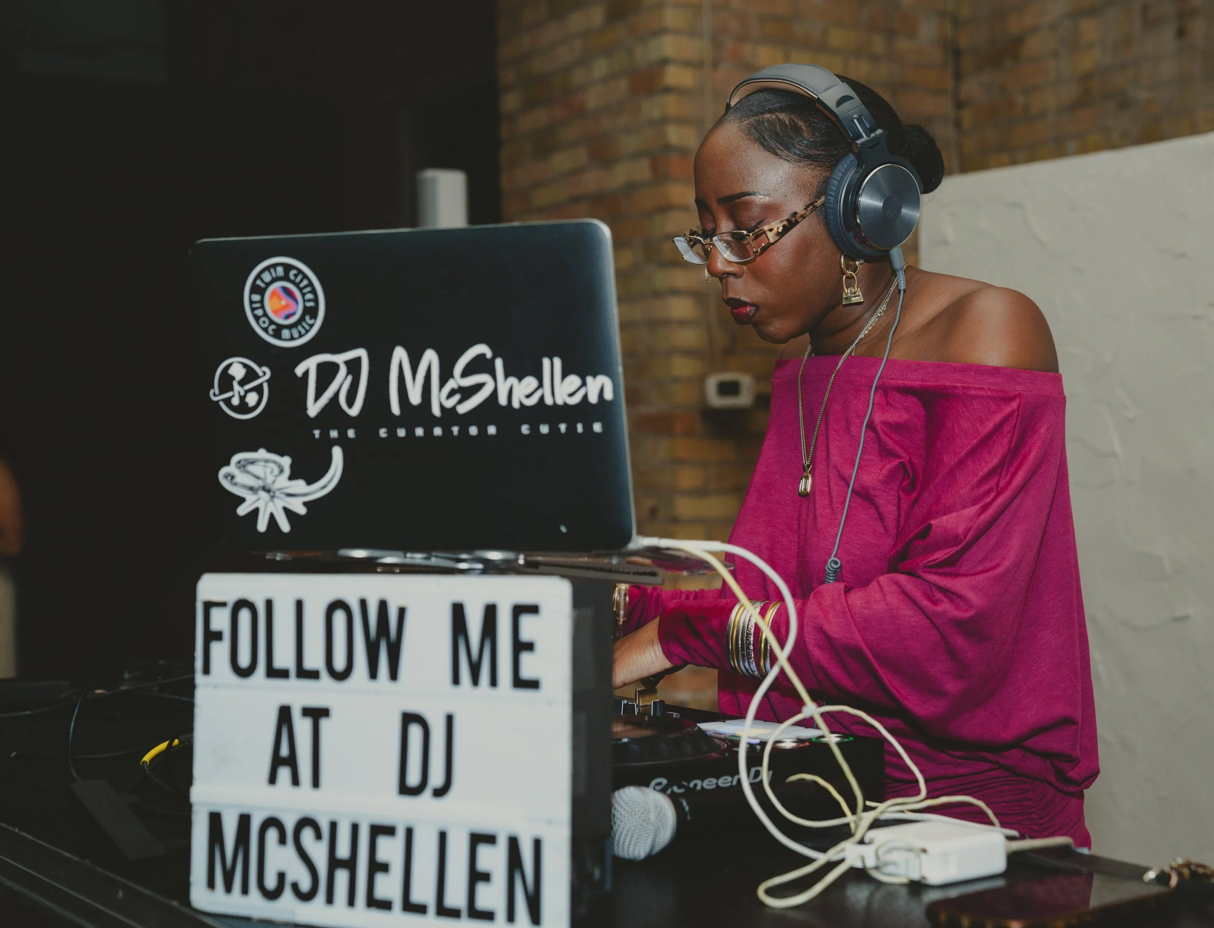 A woman with dark skin, glasses, red lipstick, and earrings wearing a pink off-shoulder top, with headphones, working on a laptop with DJ stickers and a sign that says 'FOLLOW ME AT DJ Mischel'.
