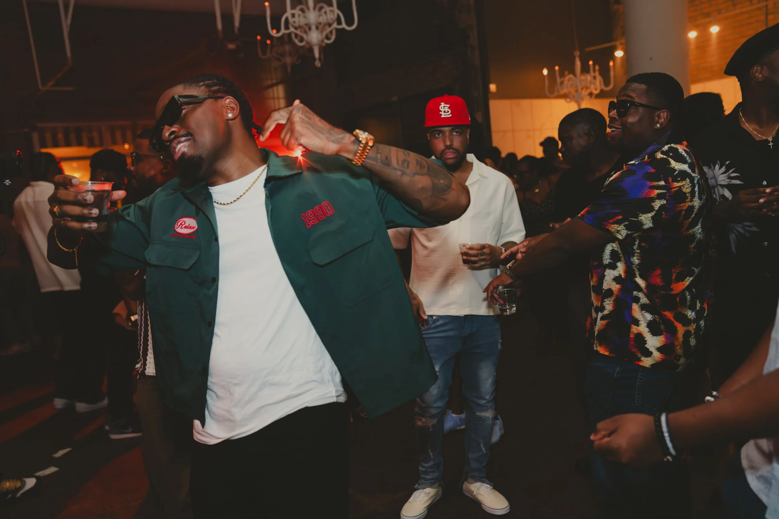 Group of people dancing and socializing at a party or nightclub with low lighting, chandeliers, and brick walls.