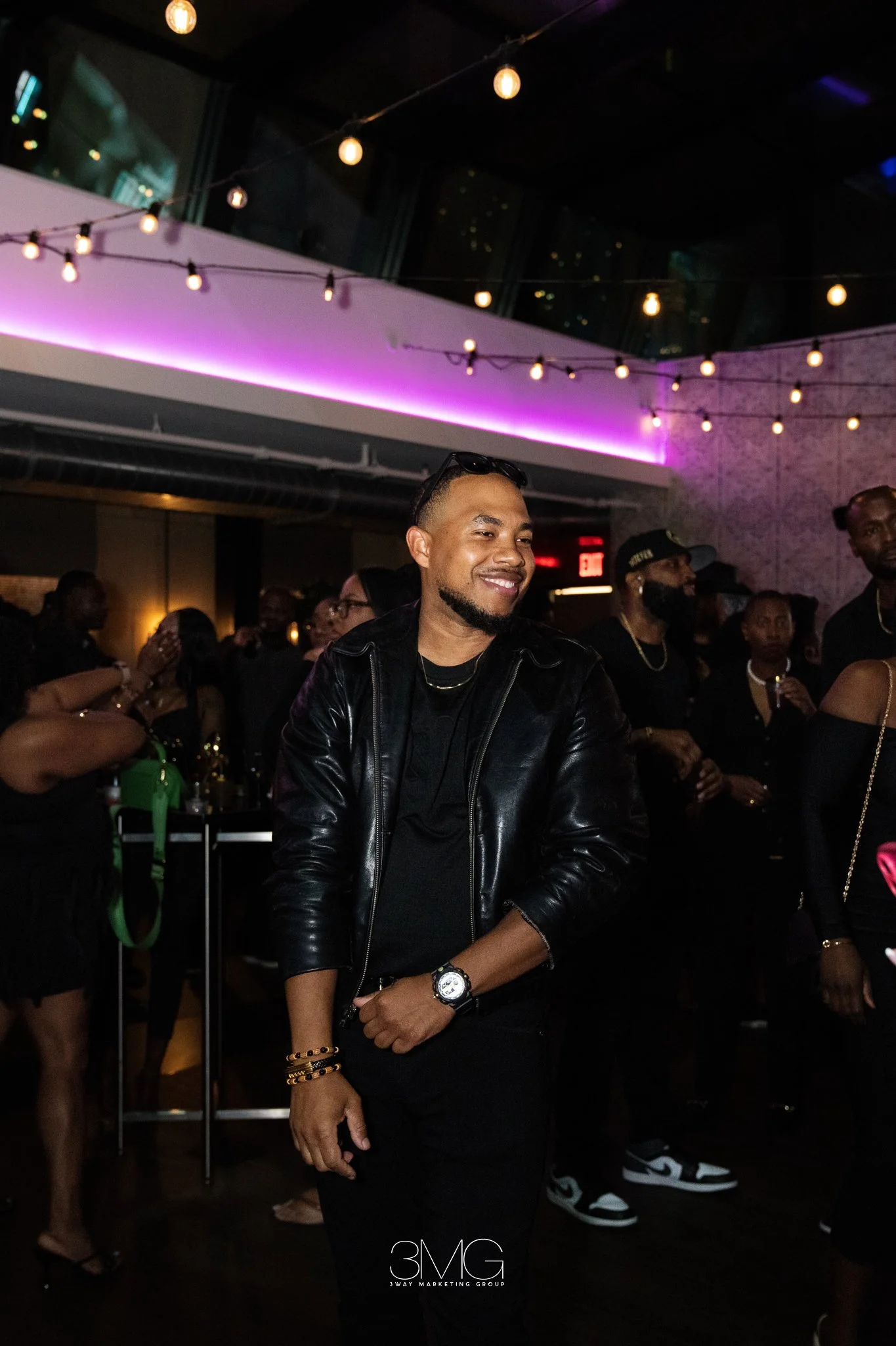 A man smiling at a party or nightclub, wearing a black leather jacket, black shirt, and jewelry, with other people in the background and string lights overhead.