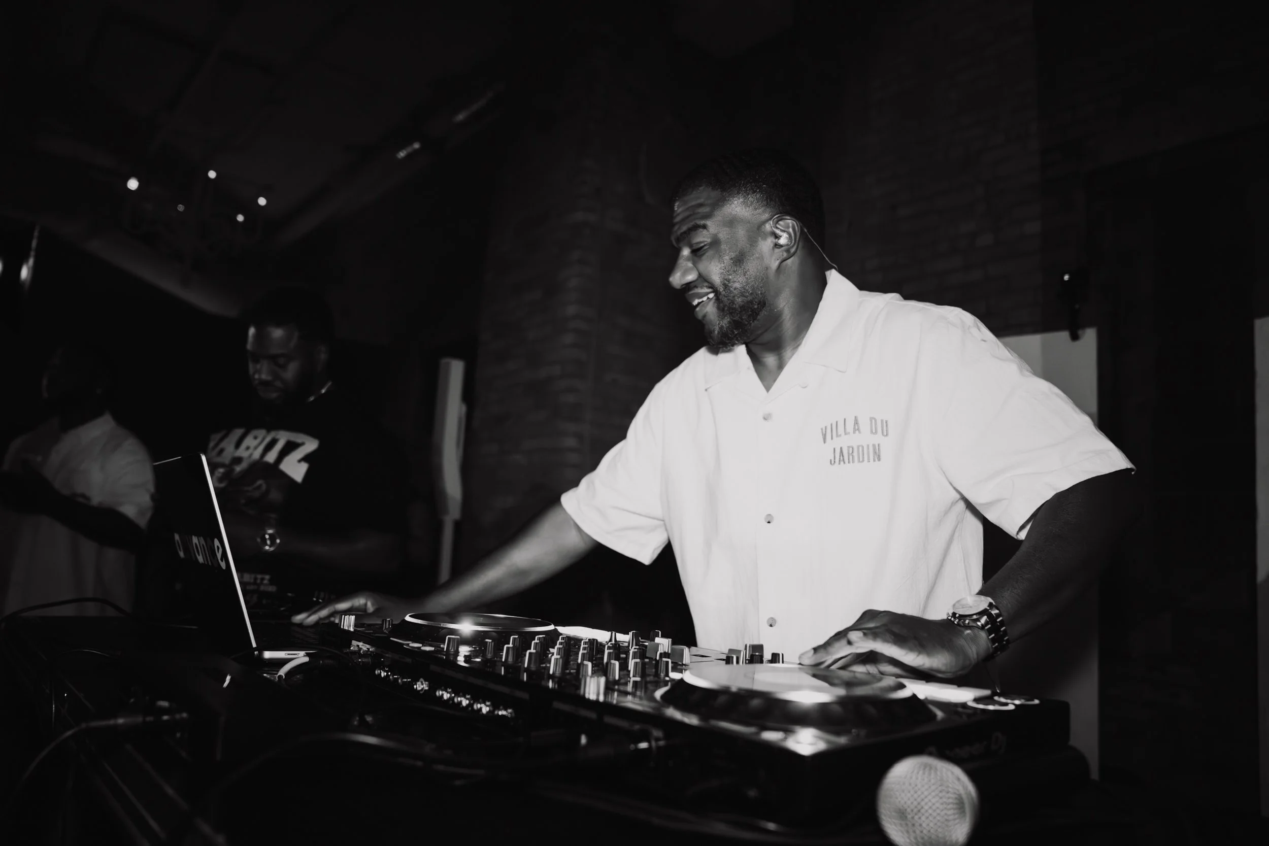 Black and white photo of a DJ smiling and working with music equipment, wearing a white shirt that says 'Villa Du Jardin'.