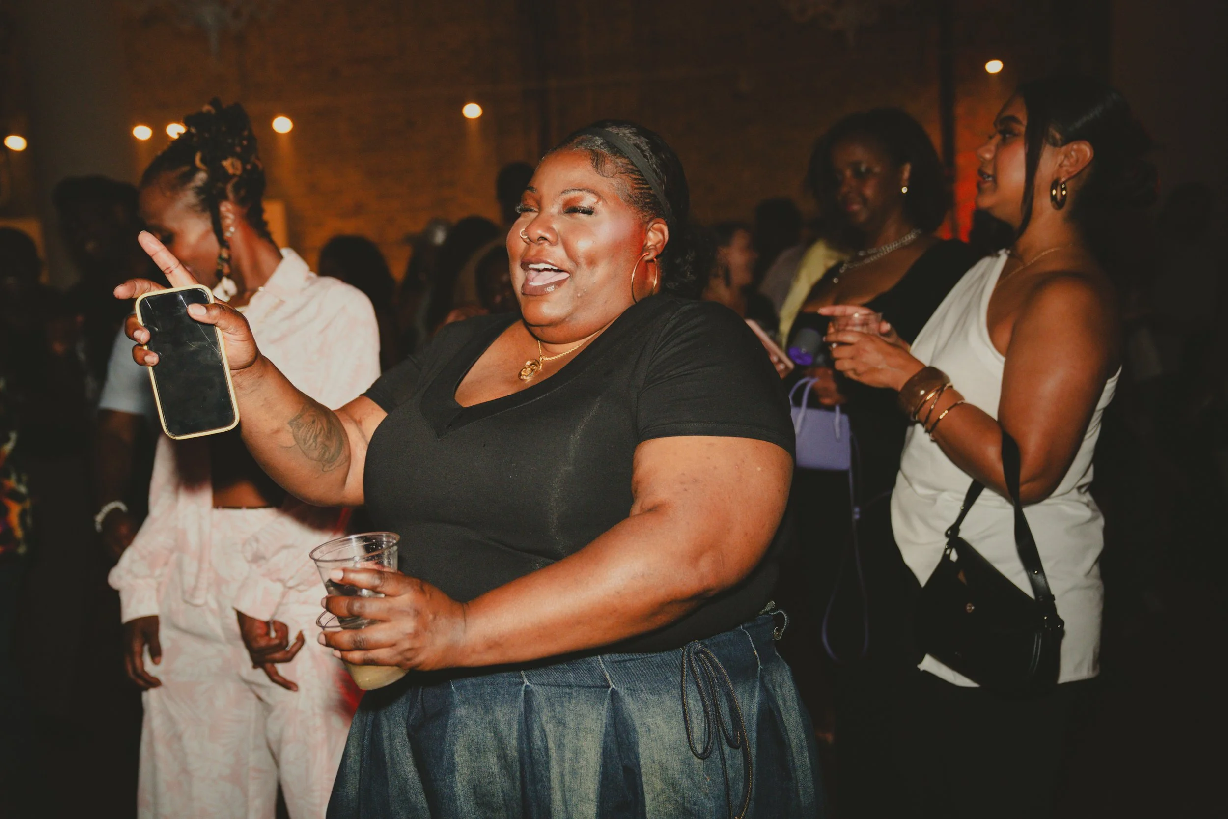 Group of women at a social gathering, smiling and enjoying the moment, one woman holding a phone and another holding a drink, in a dimly lit indoor setting.