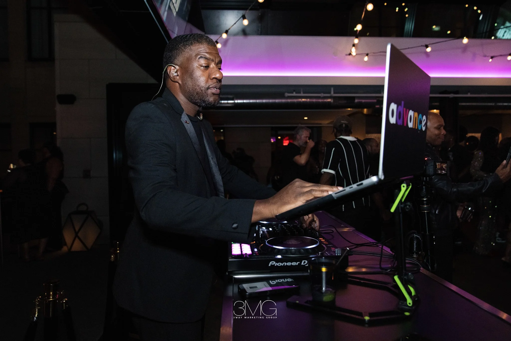 A man DJing at a party or event with a Pioneer DJ setup, surrounded by people, with dim lighting and string lights overhead.