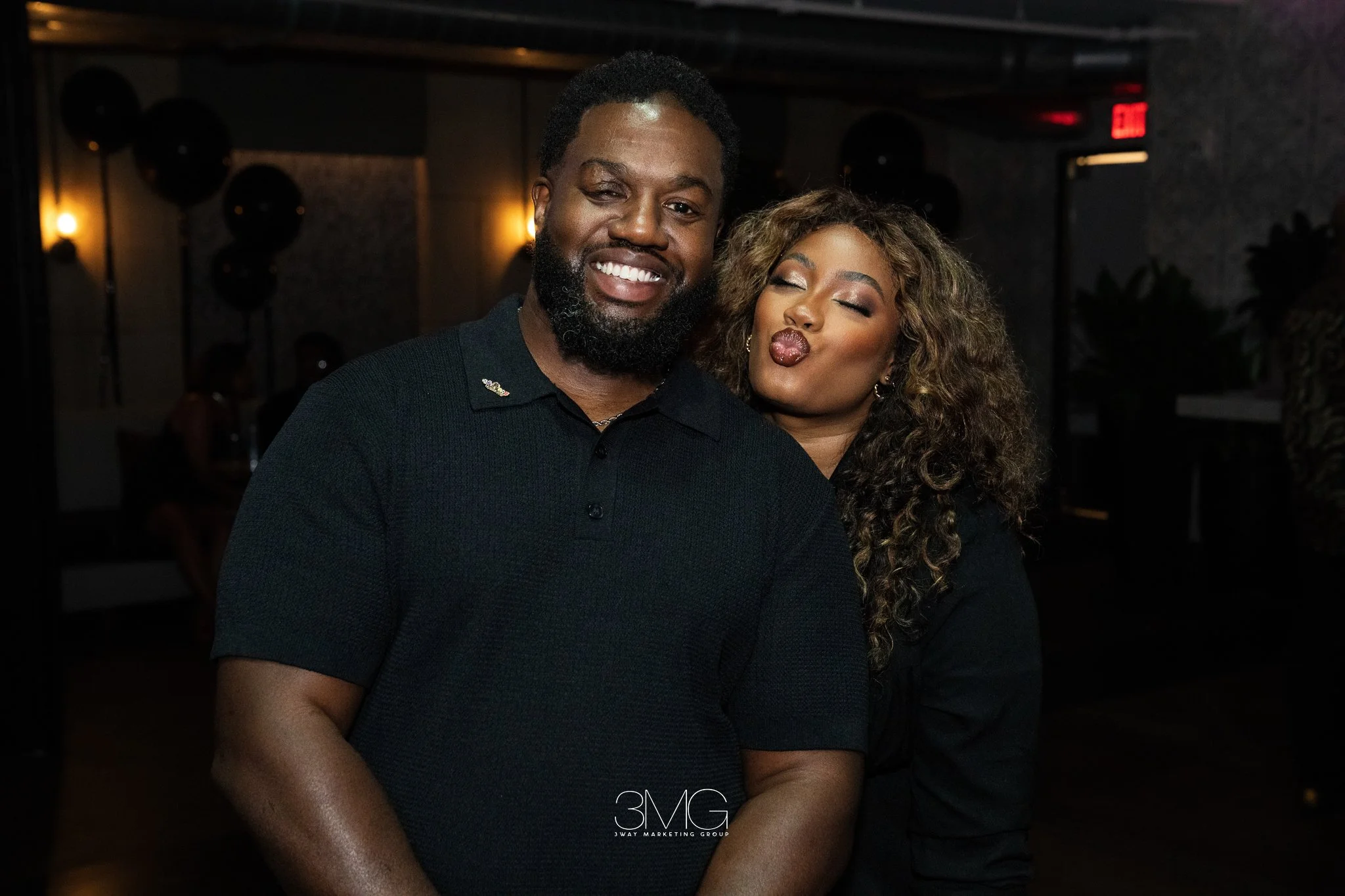 A man and woman posing for a photo at a social event. The man has a beard and is smiling broadly, while the woman is making a kiss face with her eyes closed, and has curly, light brown hair.