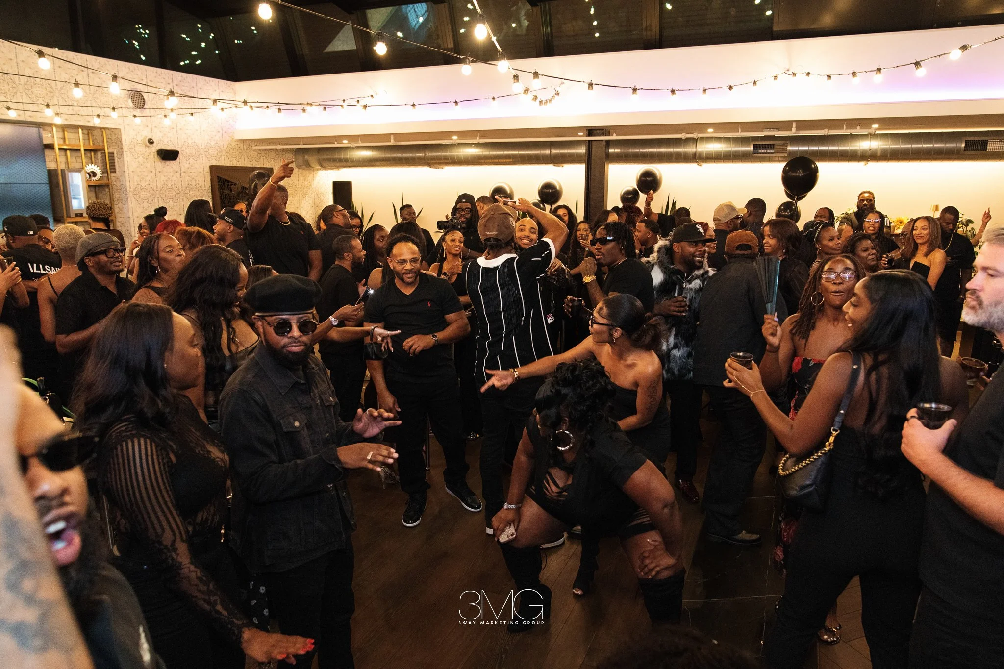 A large group of diverse people dancing and socializing at an indoor party under string lights, with some wearing sunglasses and enjoying drinks.