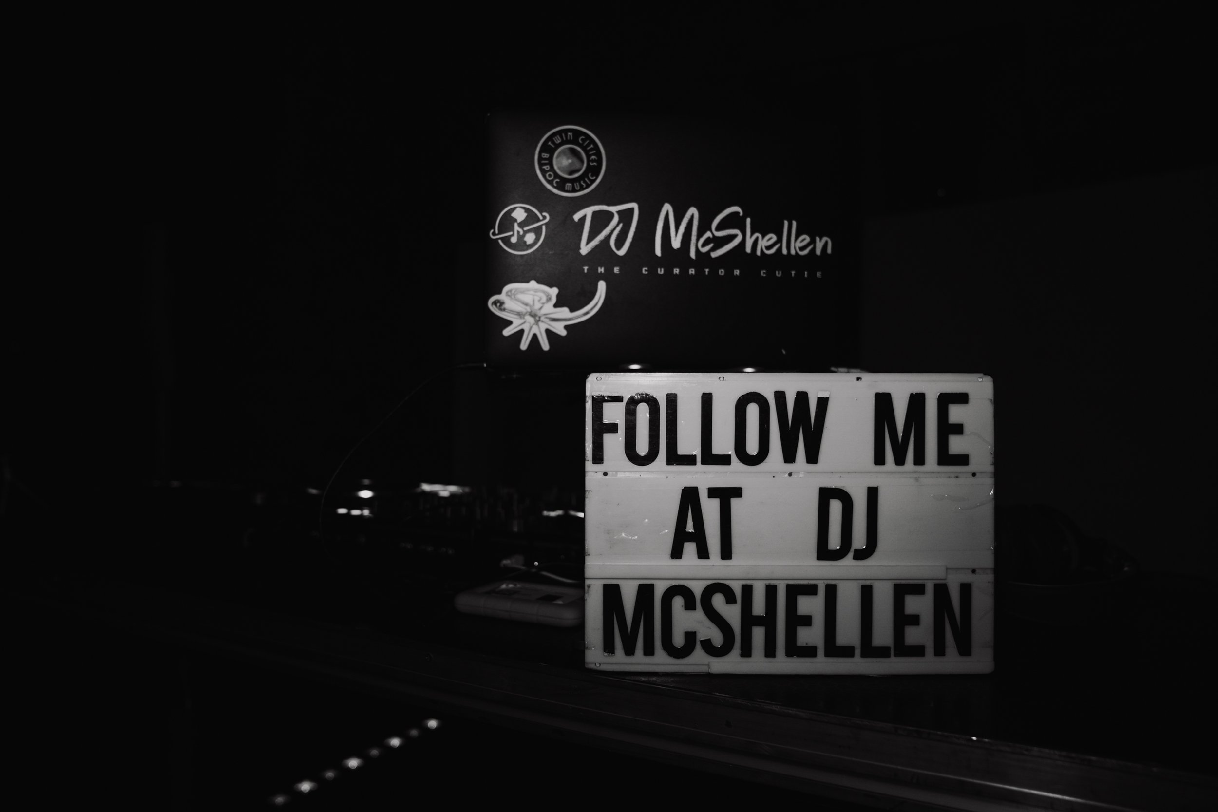 A black-and-white photo of a sign that reads 'FOLLOW ME AT DJ MCSHELEN' and a smaller sign above it featuring two illustrations and text about Twin Cities BIPOC music and DJ McShelly.