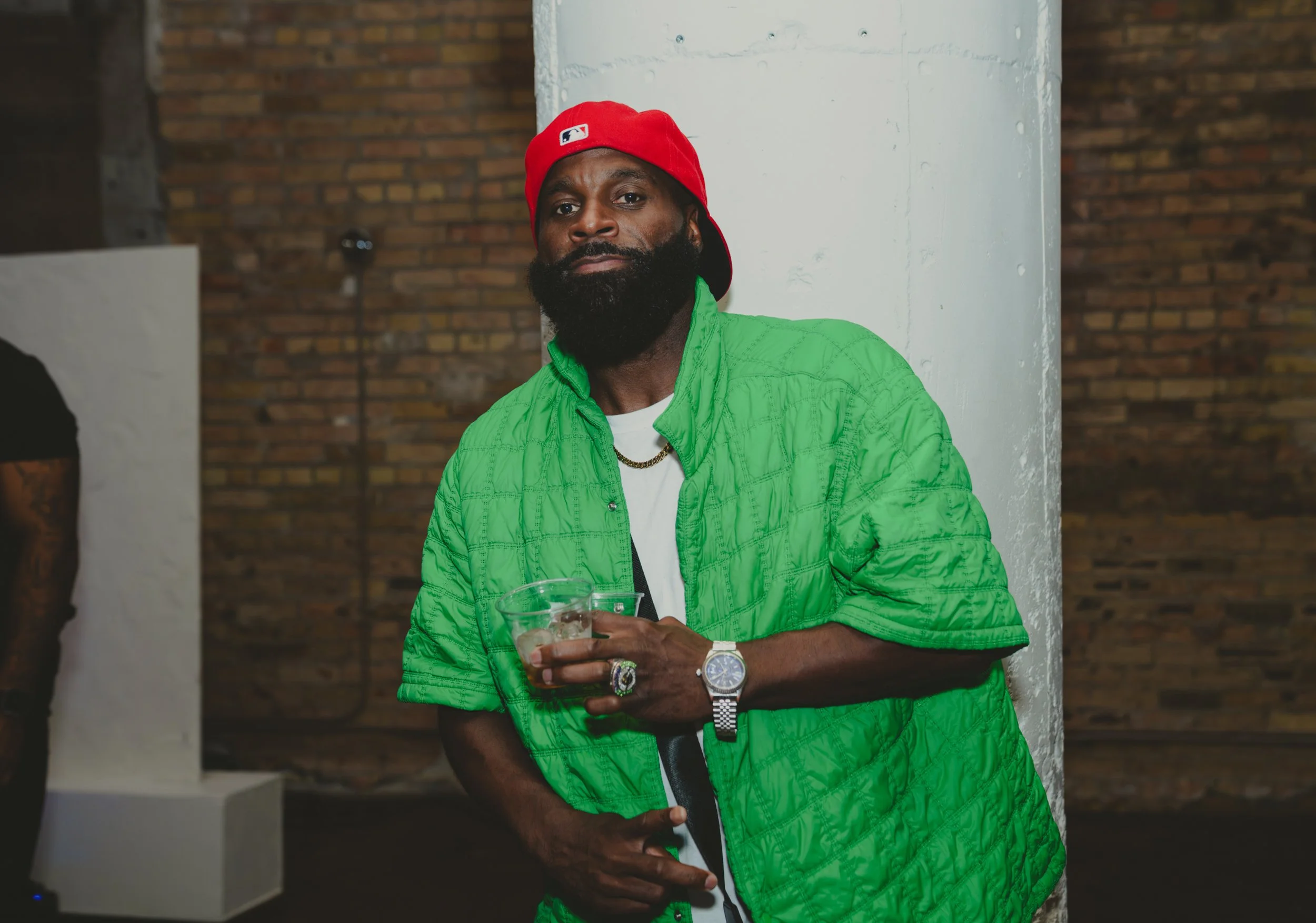 A man with a dark beard and mustache, wearing a red baseball cap backwards, a green jacket, white shirt, and jewelry, holding a transparent cup with a beverage, standing indoors against a background of brick wall and white pillar.