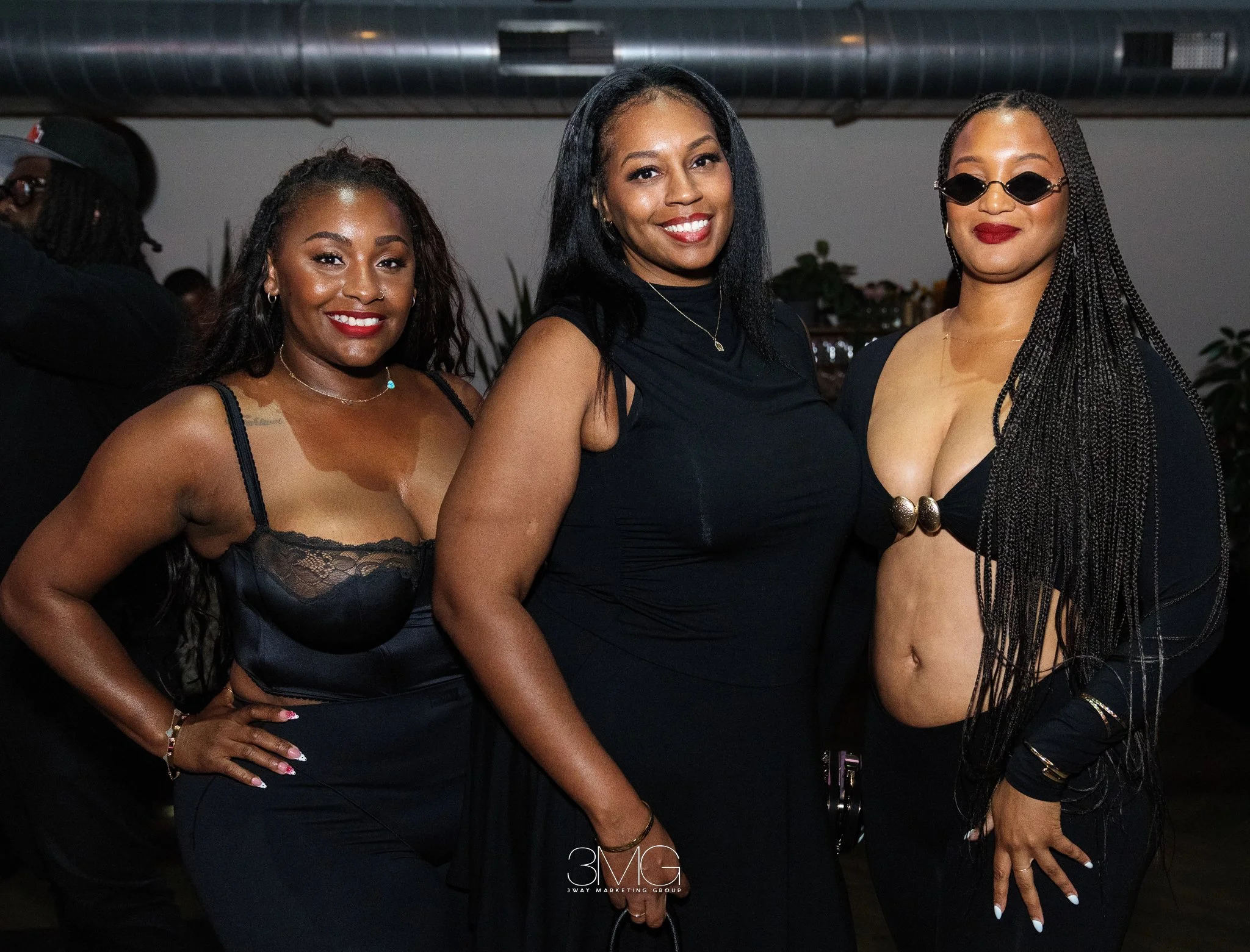 Three women smiling at a social event, dressed in black attire, standing close together in a dimly lit indoor setting.