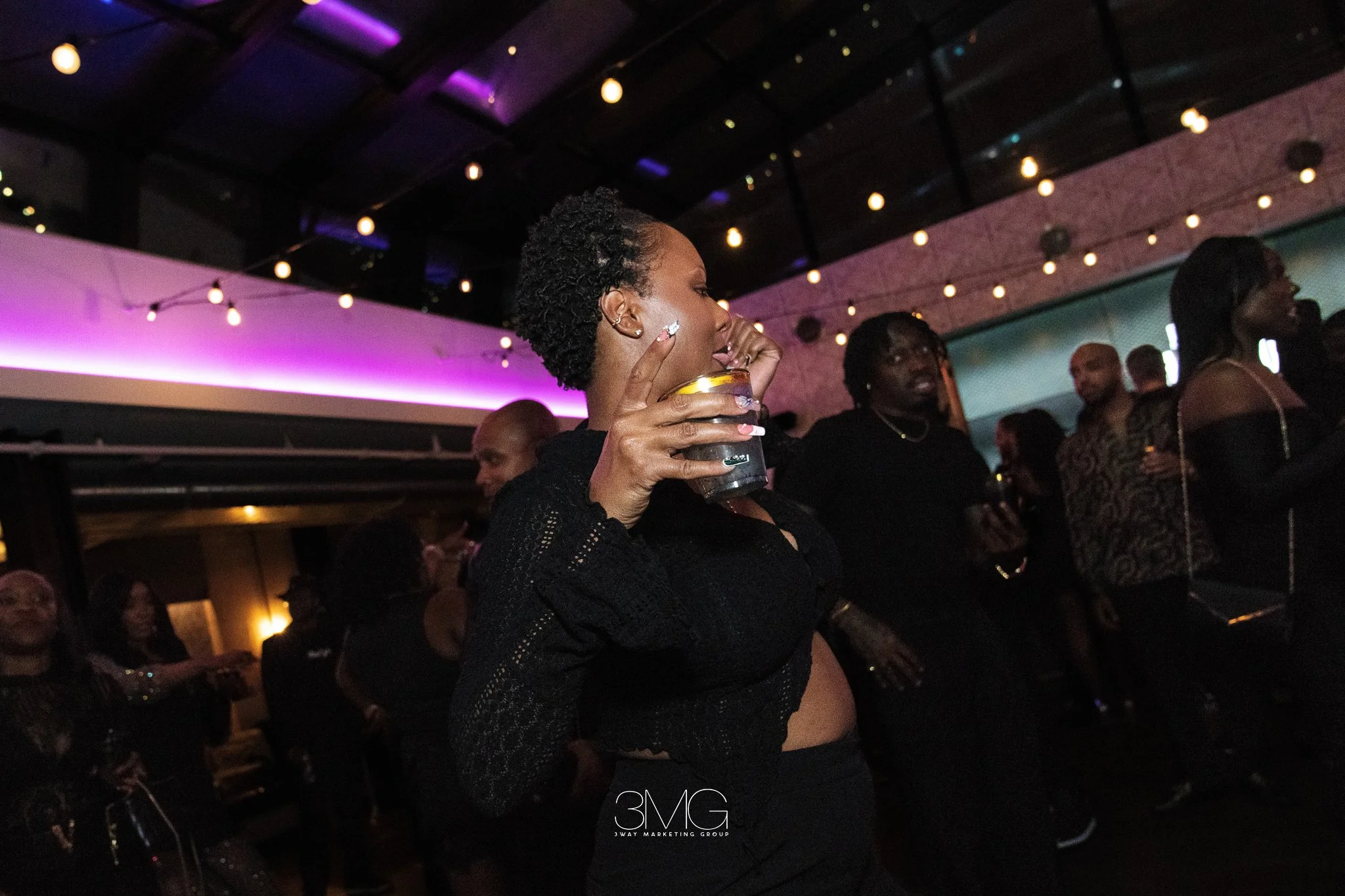 A woman with short curly hair holding a drink at a dimly lit party or nightclub. Several other people are visible in the background. String lights and purple lighting are seen on the ceiling.