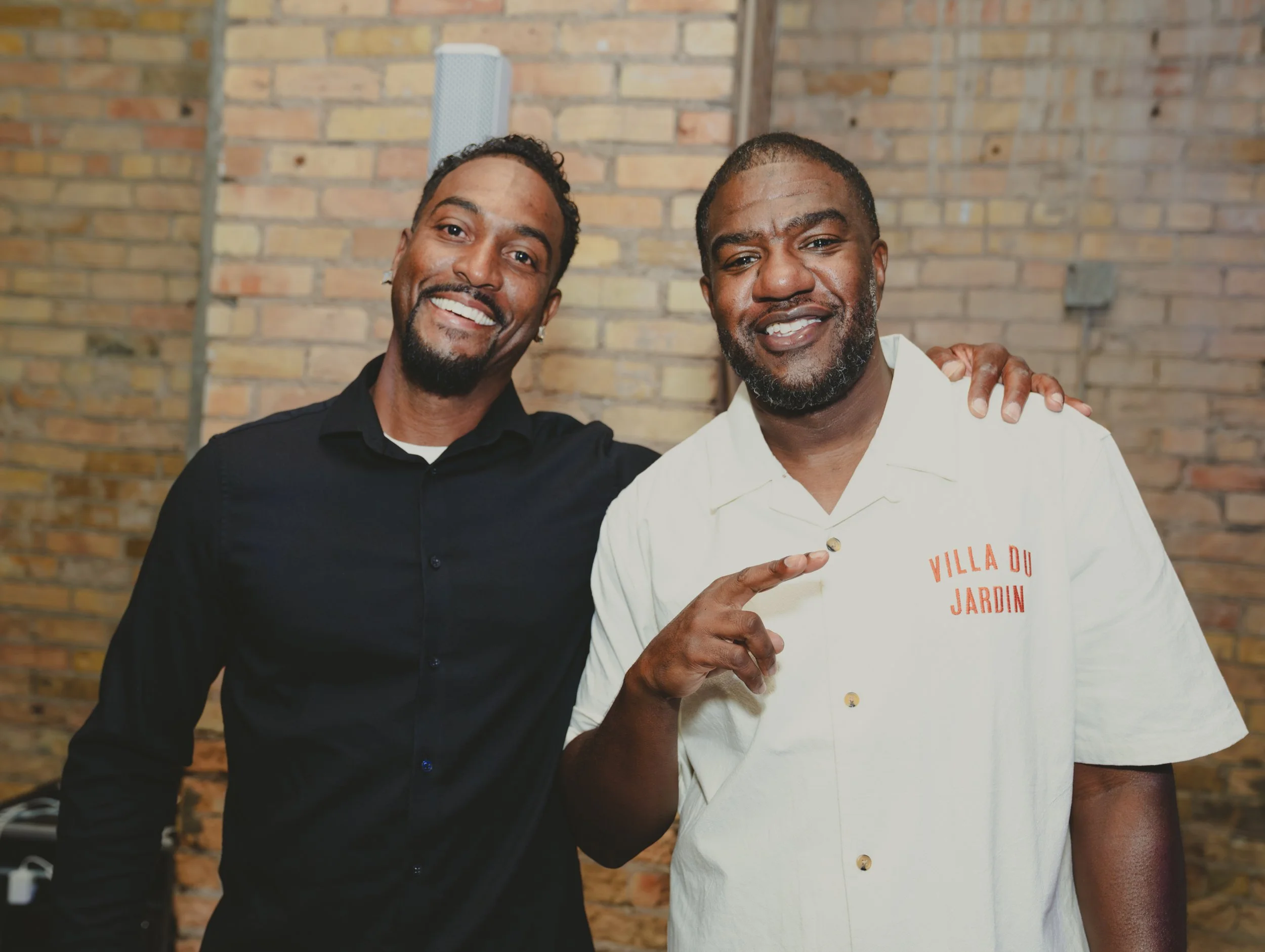 Two smiling men posing together in front of a brick wall, one wearing a black shirt and the other wearing a white shirt with orange text.