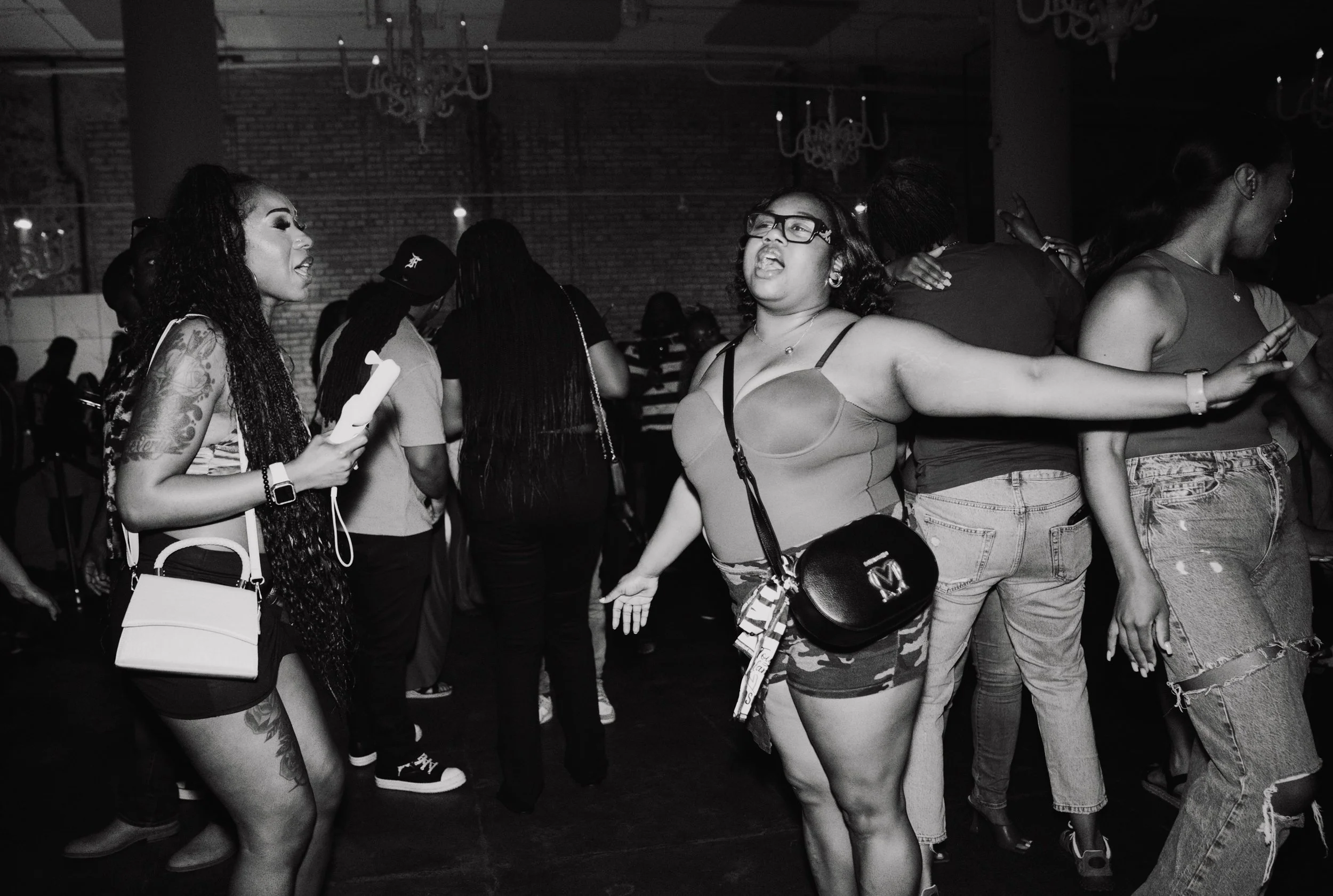 A group of young women dancing and socializing at a party or club, with some wearing casual clothes, in a dimly lit indoor venue with chandeliers. The photo is in black and white.