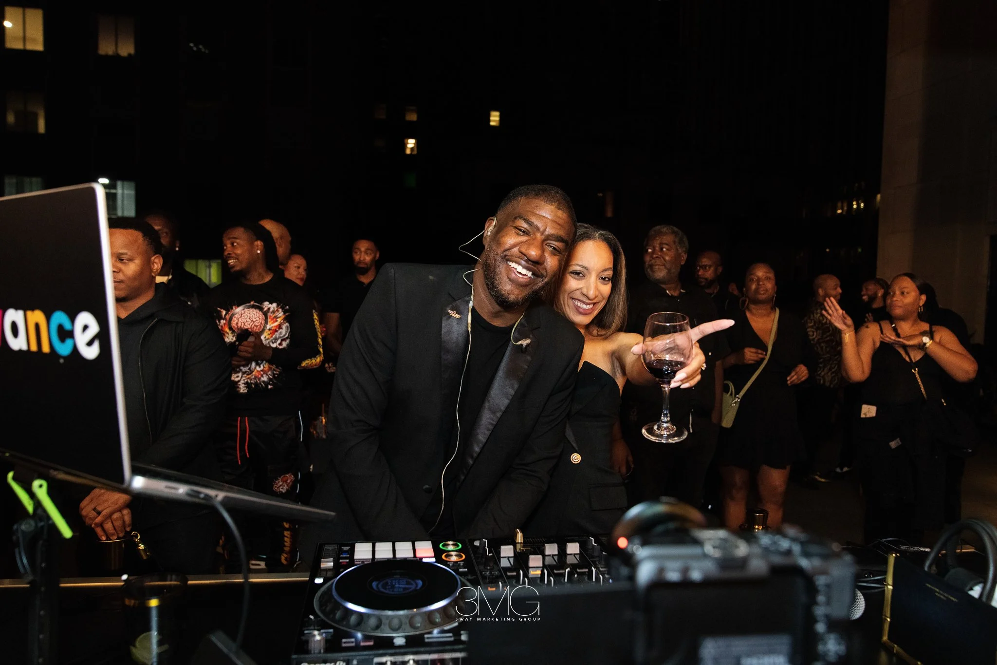 A man and woman smiling and enjoying drinks at a party or event, with other people in the background; a DJ setup with equipment is visible in the foreground.