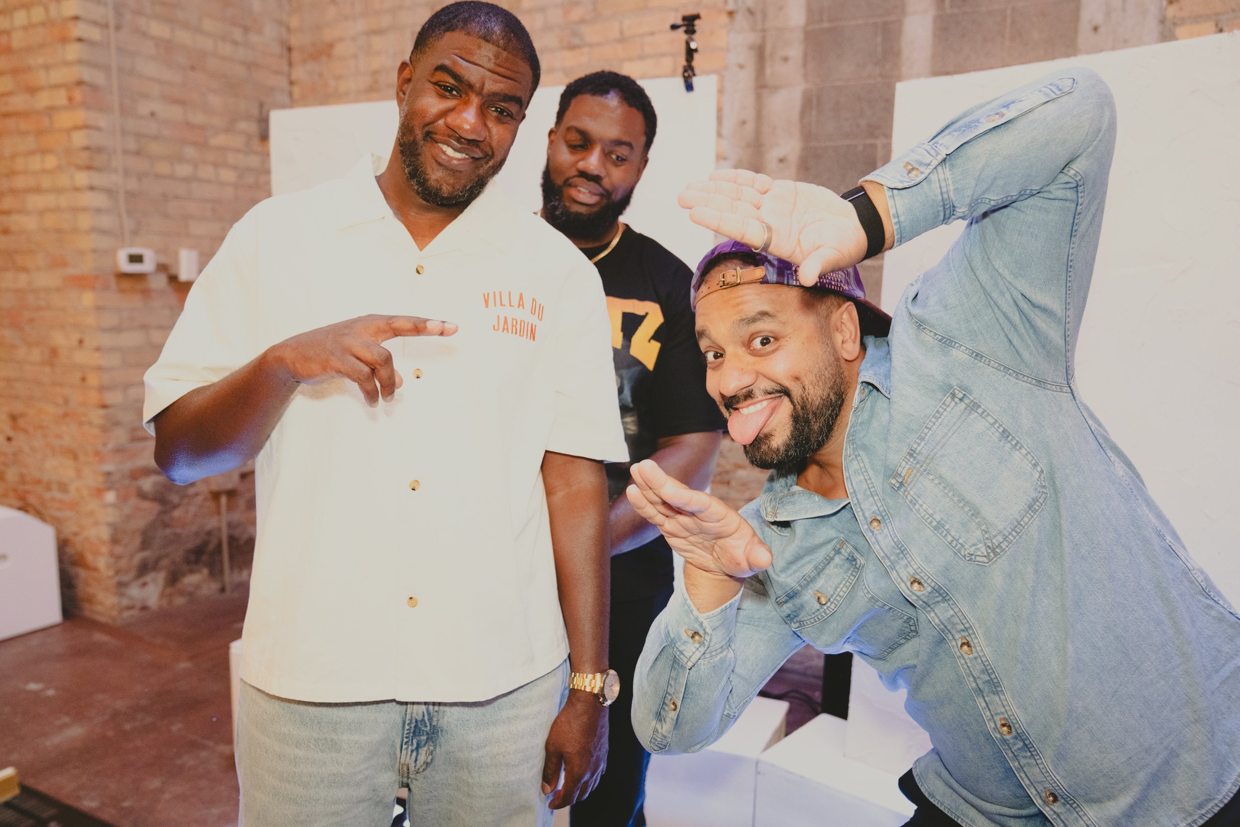 Three men are posing playfully in an indoor space with exposed brick walls. The man in front is wearing a denim jacket and a baseball cap backward, sticking out his tongue. The man on the left, dressed in a white shirt with gold buttons and 'Villa Du