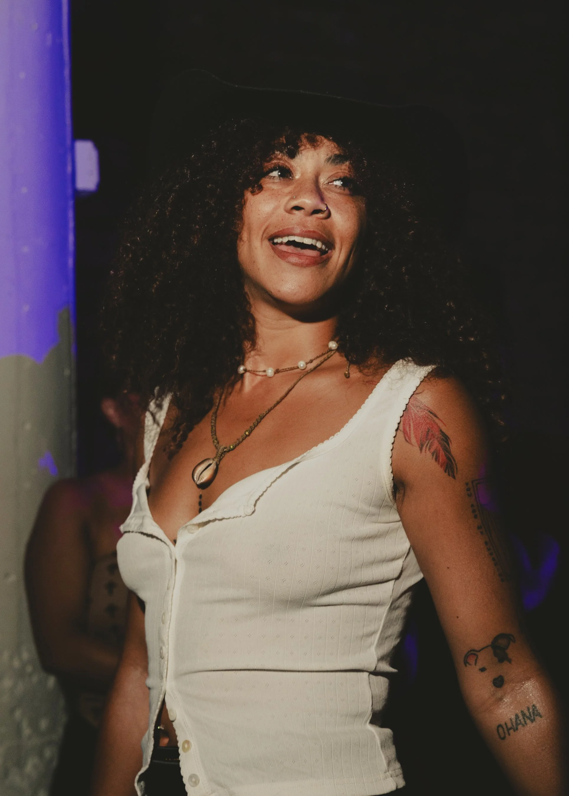 A woman with curly hair wearing a black hat, a white sleeveless top, layered necklaces, and tattoos on her arm.