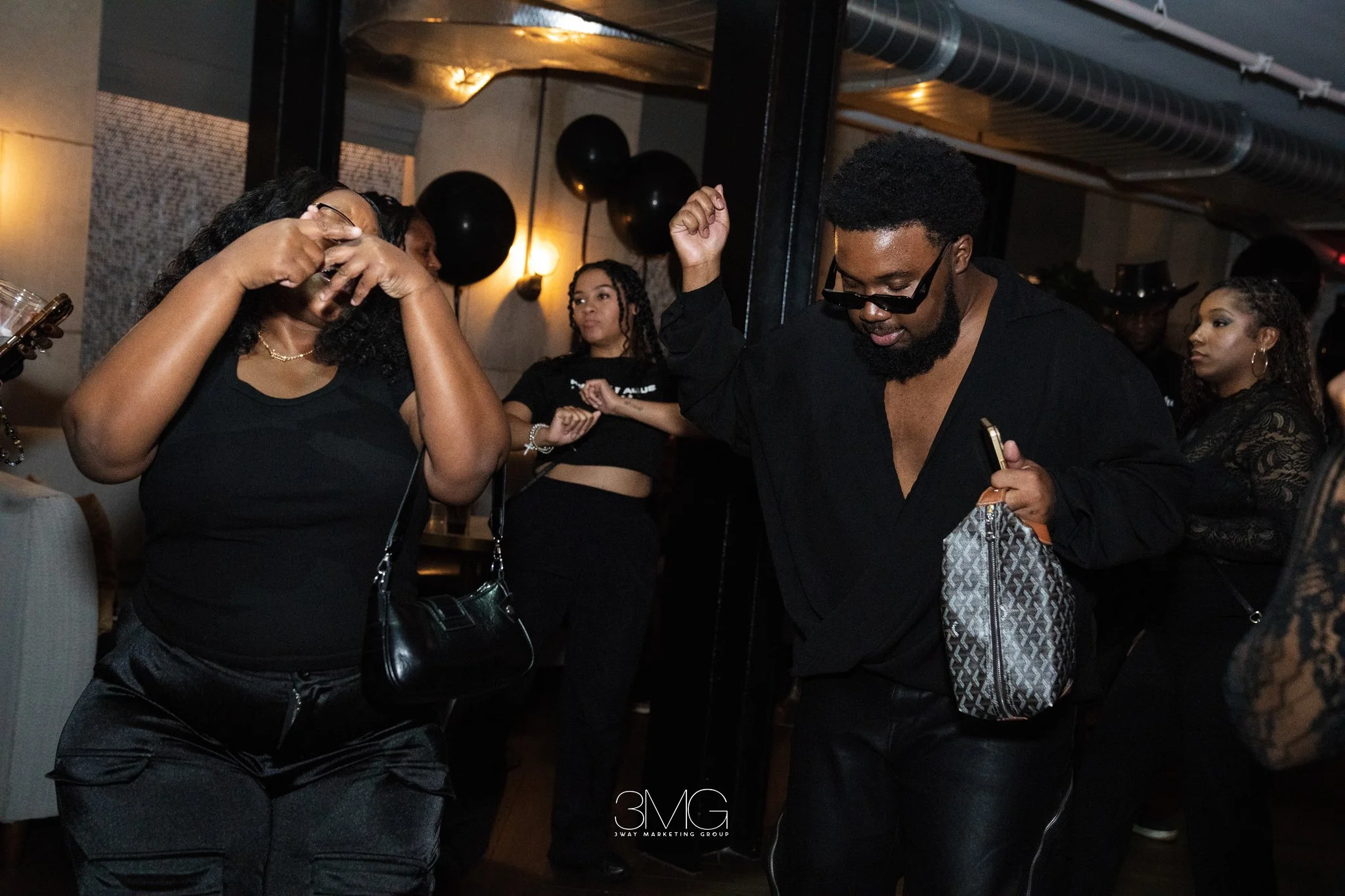 People dancing and socializing at a party in a dimly lit room with black balloons and warm yellow lighting.