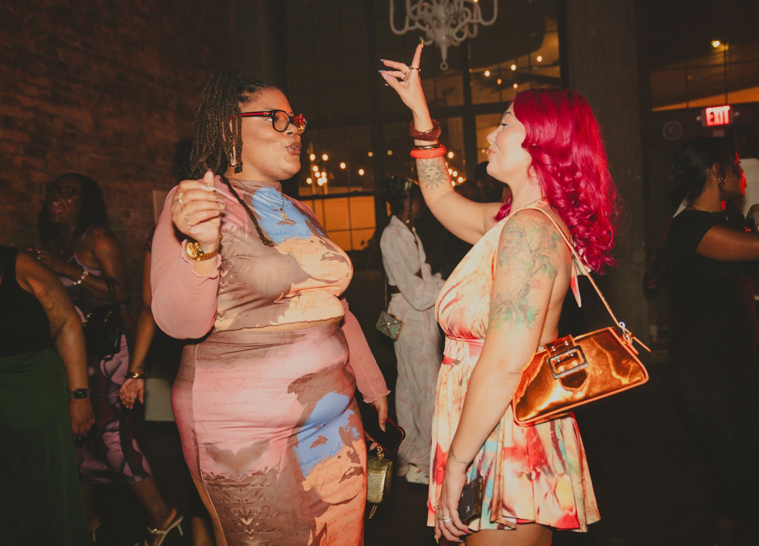 Two women with colorful dresses dancing and talking in a dimly lit party or club setting, with other people in the background.