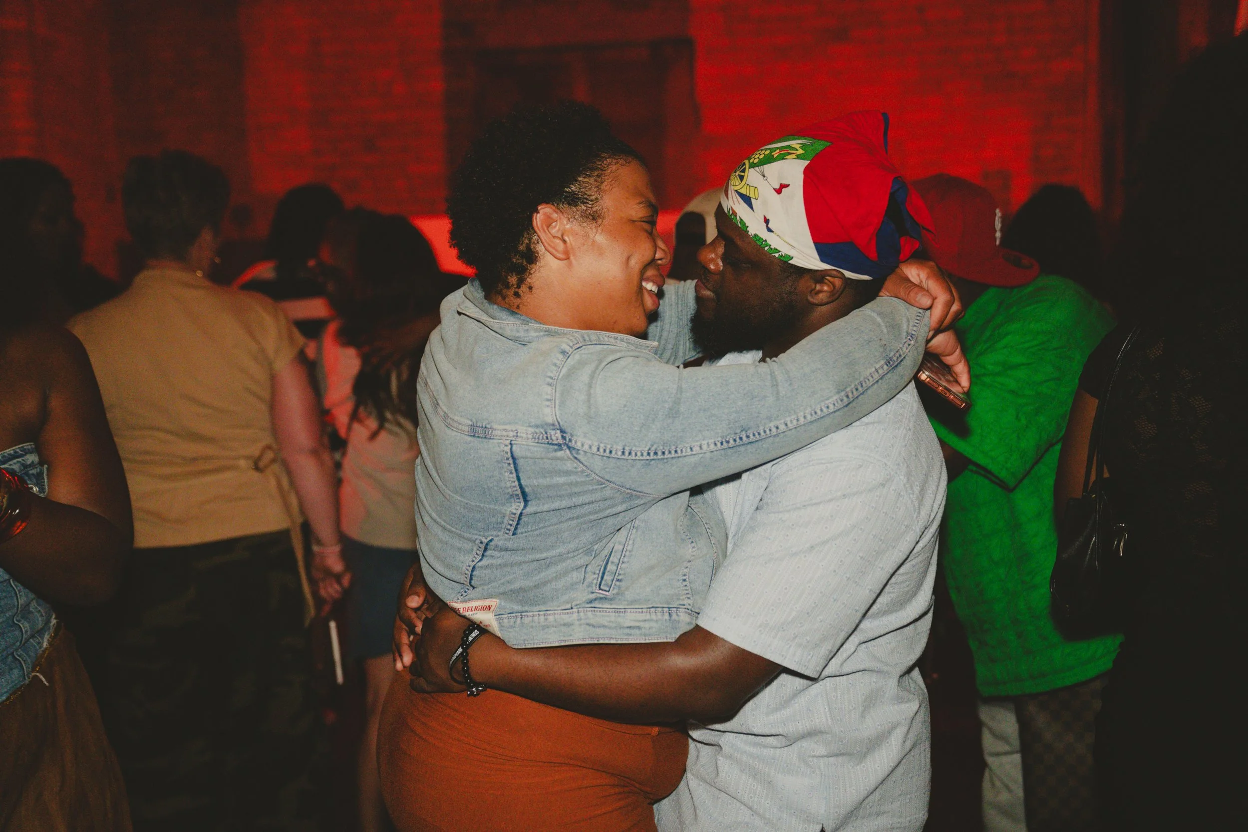 A woman and a man are dancing closely; she has short curly hair and is smiling, wearing a denim jacket and orange pants, while he is wearing a colorful headscarf and a light-colored shirt. The background shows other people in a dimly lit room, possib