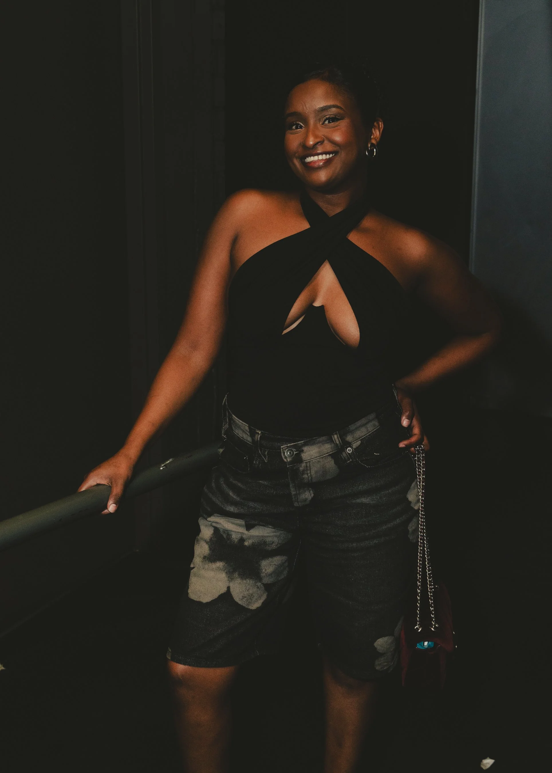 A smiling woman with short hair wearing a black halter top with cutouts, camouflage shorts, and carrying a small purse with a chain strap, standing in a dark environment.