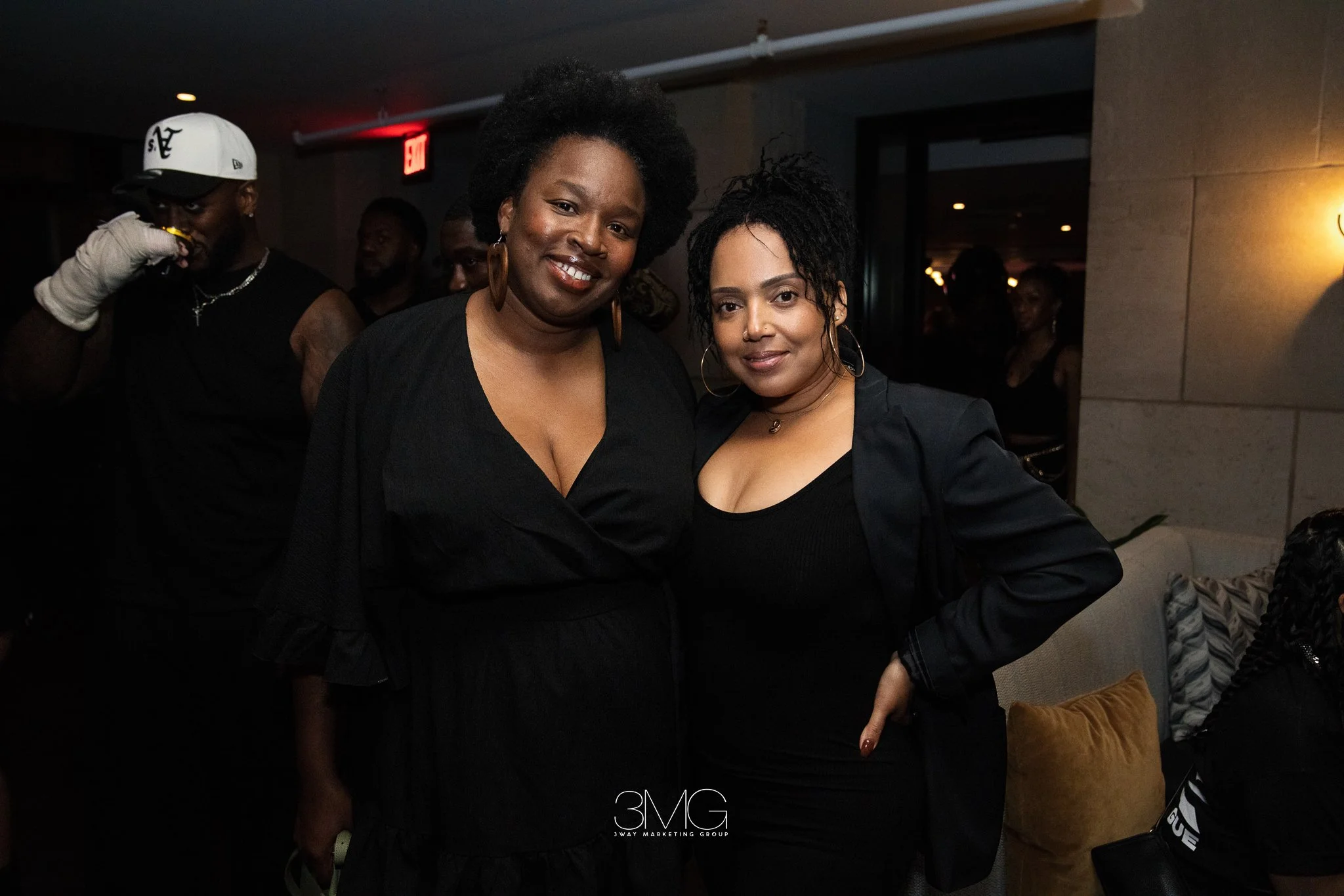 Two women smiling at the camera in a dimly lit indoor setting, with several other people in the background. One woman has a natural afro hairstyle, and the other has short, textured hair; both are wearing black tops. The setting appears to be a socia