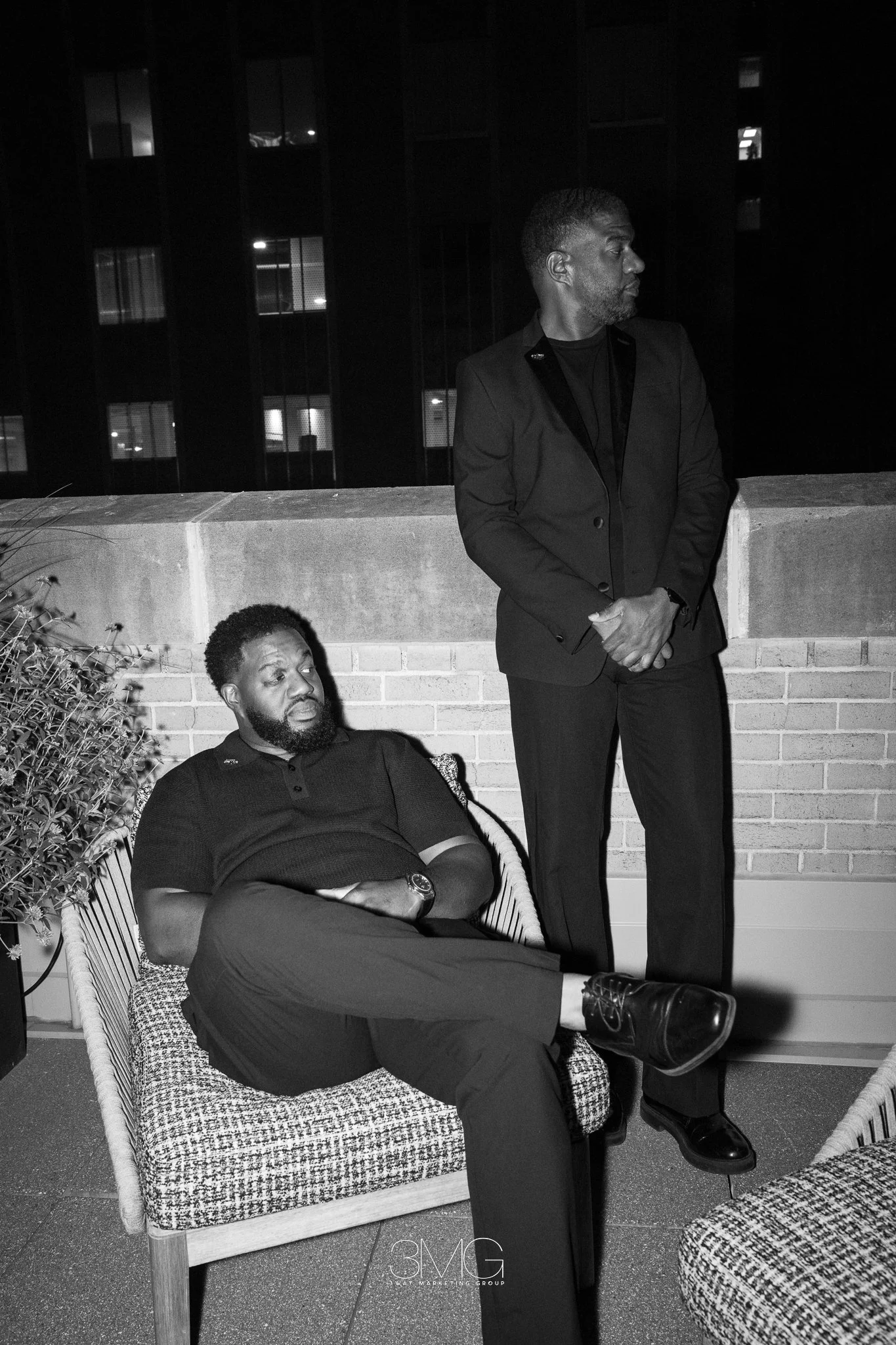 A man in a suit standing on a rooftop balcony at night, looking to his right, with another man with a beard sitting on a chair, resting with his eyes closed, and a woman with curly hair sitting nearby, all surrounded by plants and a brick wall.