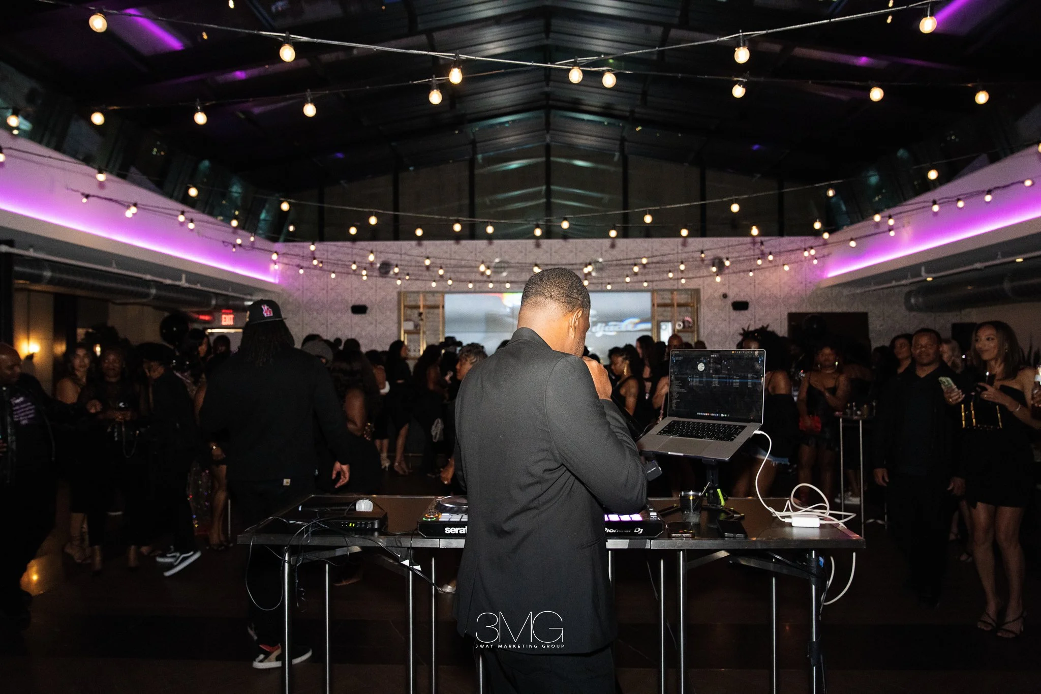 A DJ performs at a crowded indoor event with string lights and purple lighting, with people dancing and socializing.