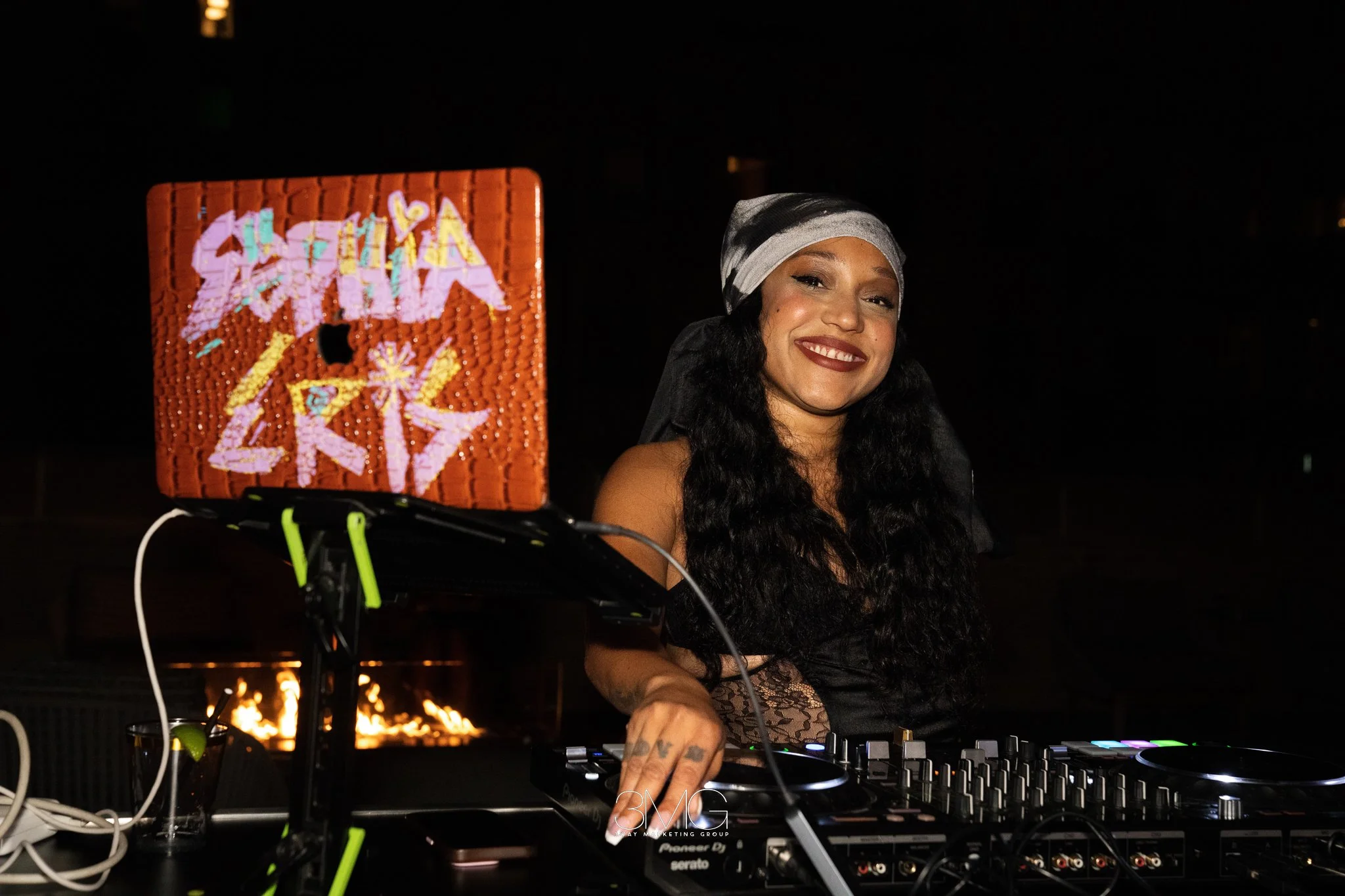 A woman with long dark curly hair wearing a silver and black headscarf, smiling while DJing at night. She is positioned beside DJ equipment with colorful buttons, and a decorated cake or box with bright graffiti-style text is visible in the foregroun
