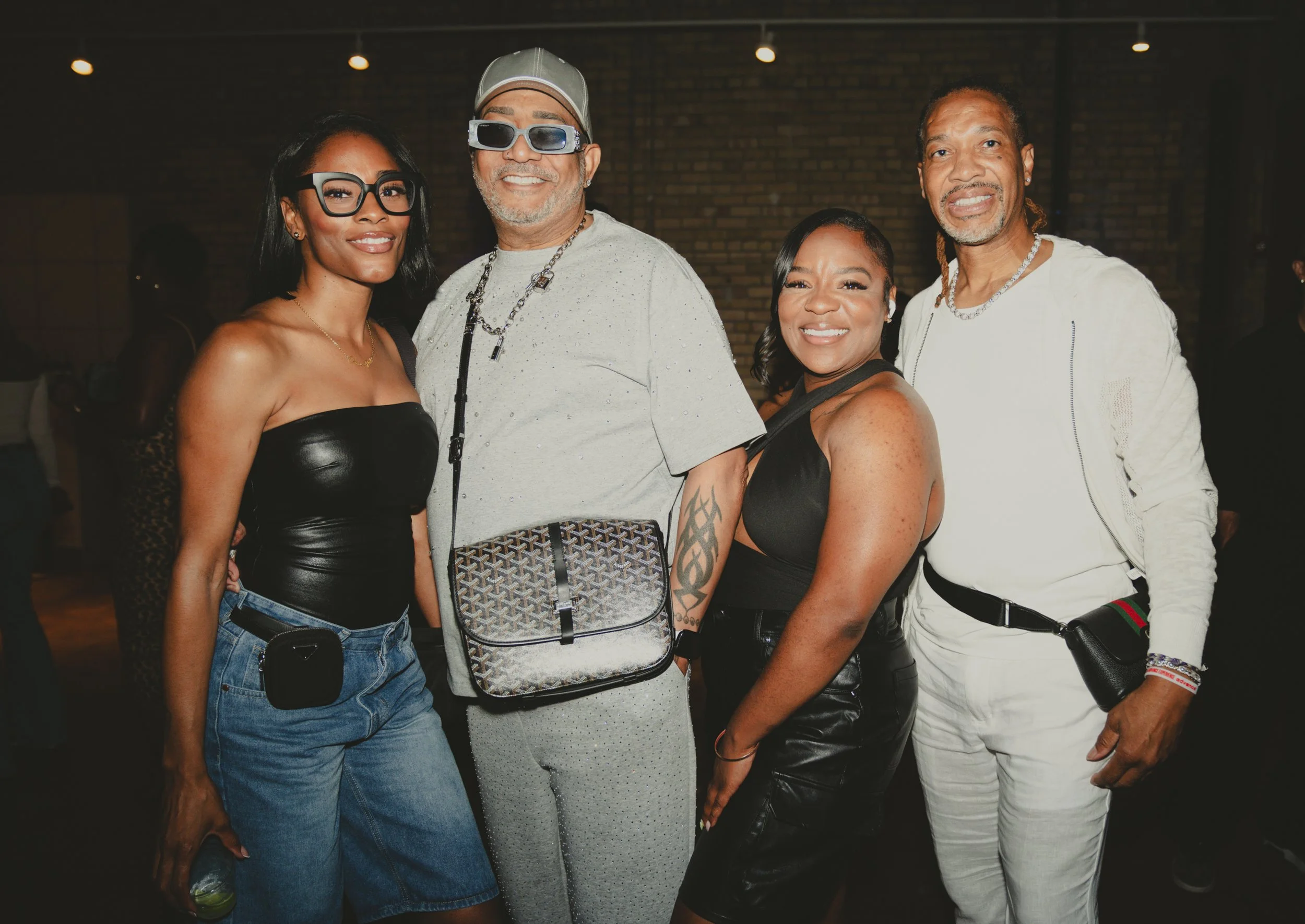 A group of four diverse adults smiling at a social event, standing close together with a brick wall in the background.