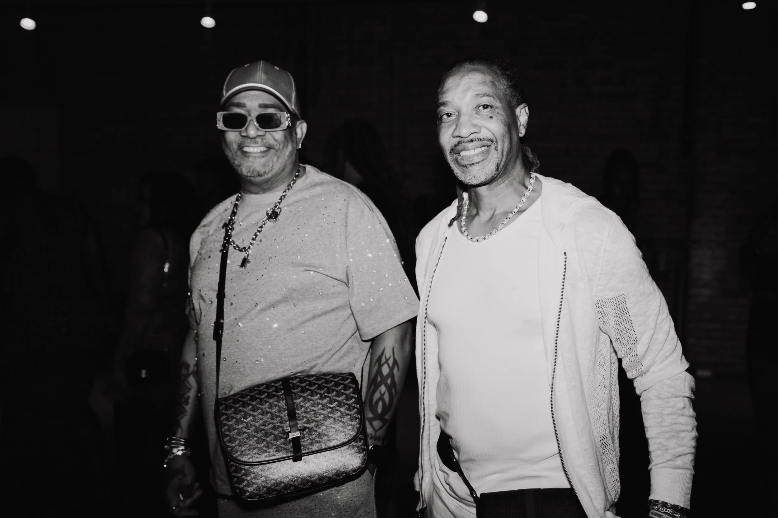 Two men smiling in a dark setting. The man on the left is wearing sunglasses, a cap, and a beaded necklace. The man on the right is wearing a light-colored jacket with a zipper and a chain necklace.