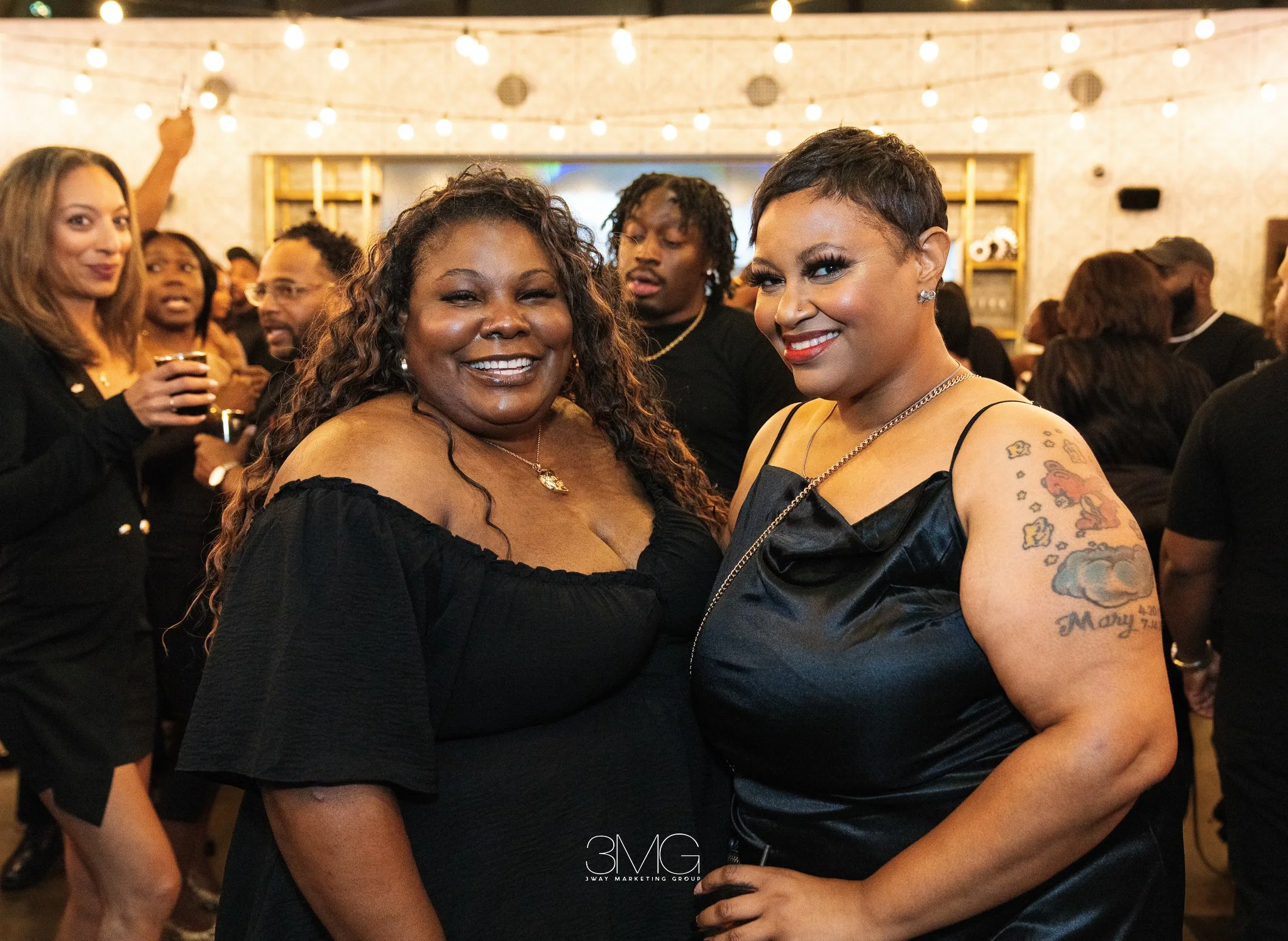 Two women smiling and posing for a photo at a social event, with other guests in the background, warm lighting, and a festive atmosphere.