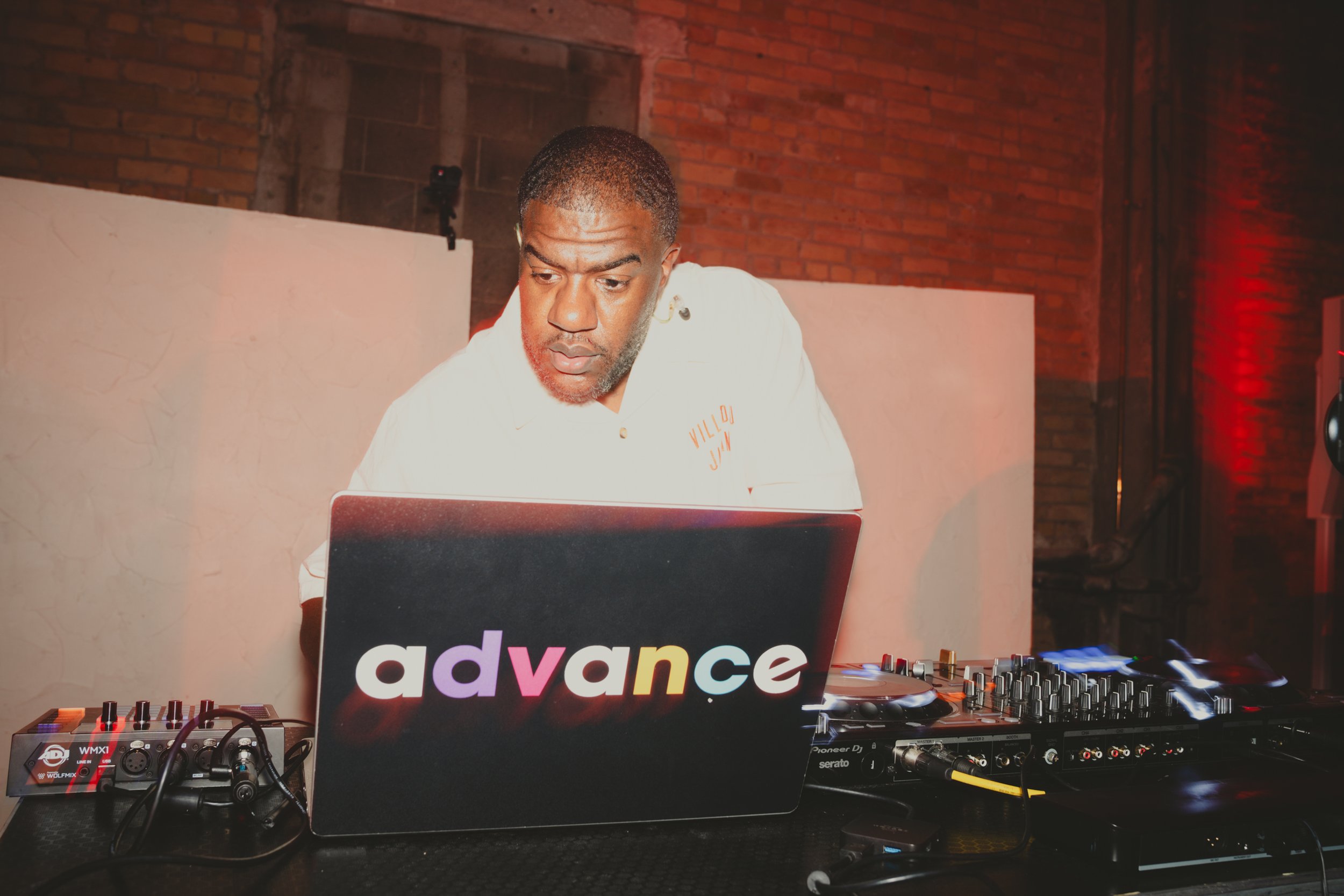 A DJ is working at a music event with a laptop that has 'advance' written on it. The DJ equipment includes a mixer and turntables, with cables connected. The background features a brick wall and dim lighting with red tones.