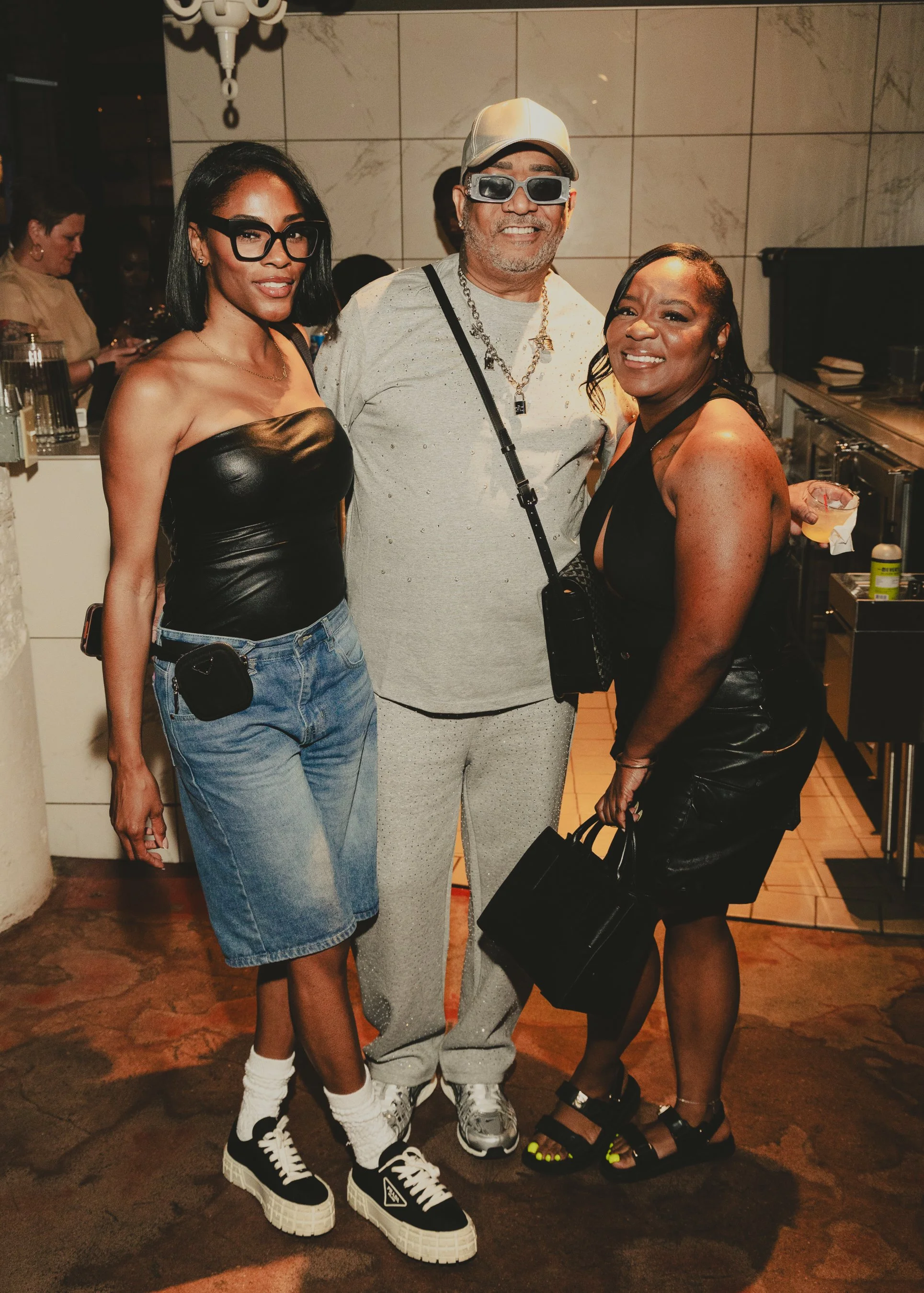 Three people standing together at a social event. The woman on the left is wearing a black leather bustier top, high-waisted denim shorts, white sneakers, chunky socks, and large black glasses. The man in the middle is wearing a light gray shirt with