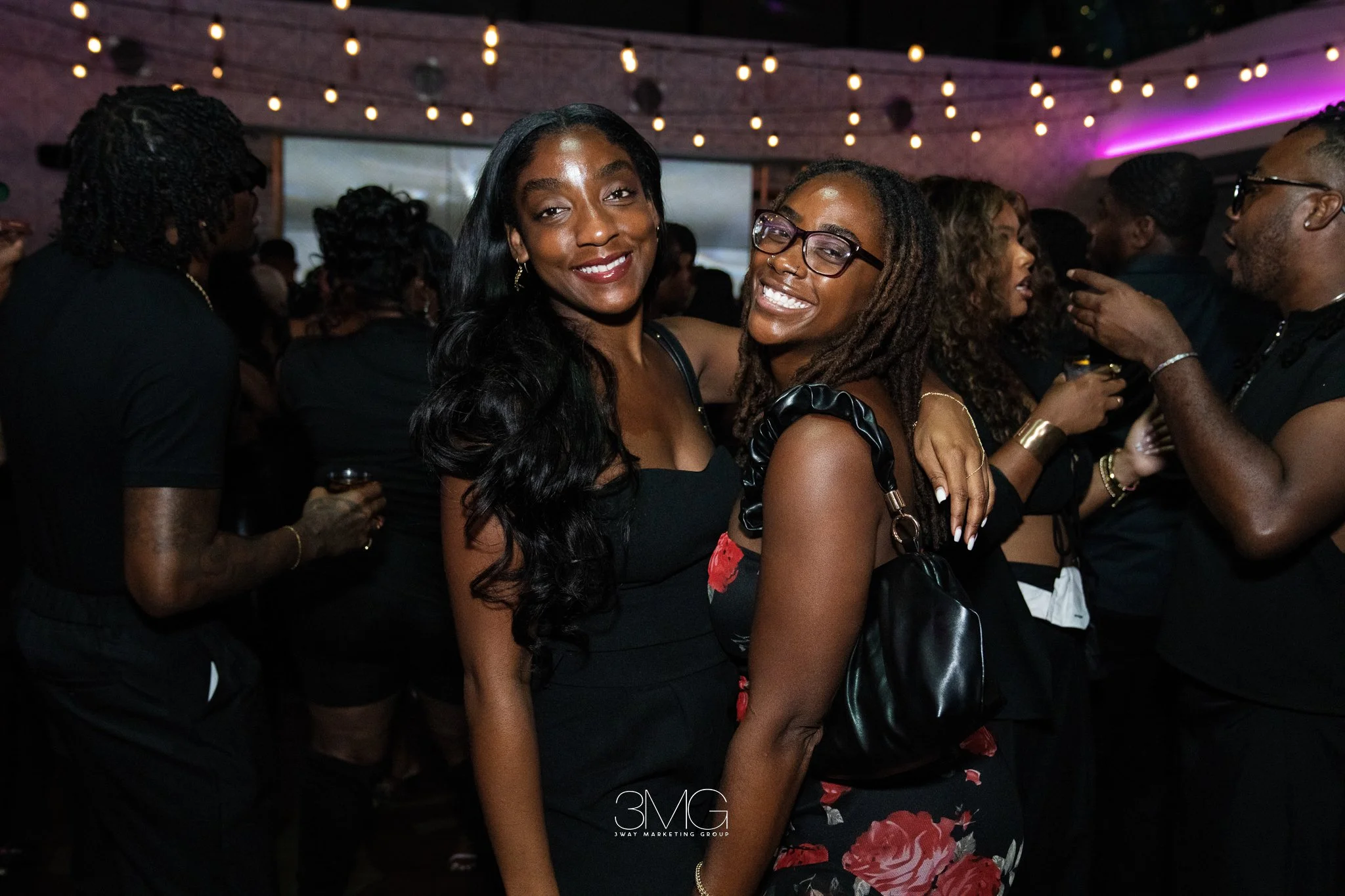 Two women with dark hair and glasses smiling at a social event, surrounded by other people under hanging string lights in a dimly lit venue.