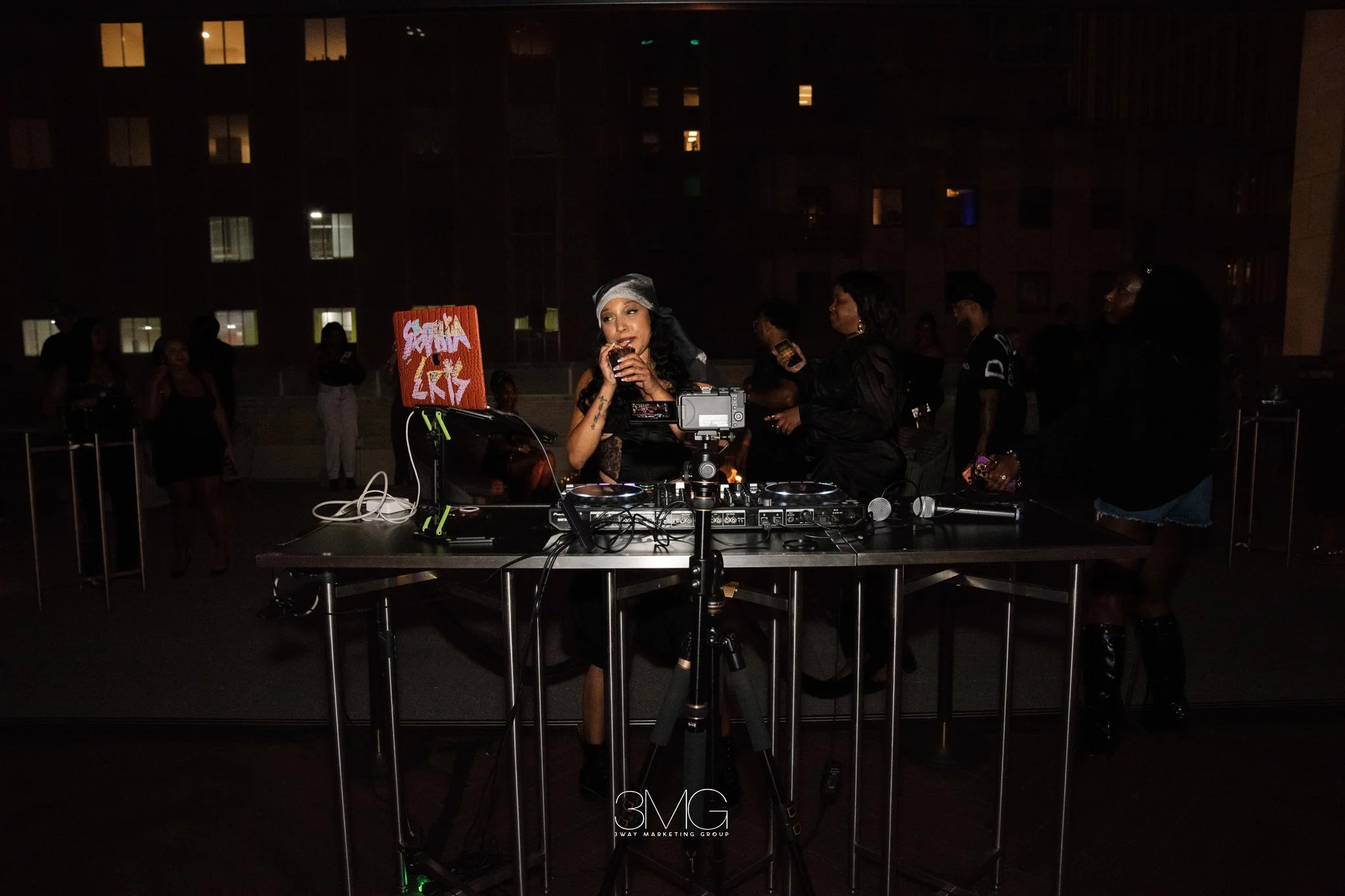 Nighttime outdoor party with a female DJ wearing a head scarf, standing behind DJ equipment and a laptop with graffiti art, surrounded by people socializing and taking photos; city buildings with illuminated windows in the background.