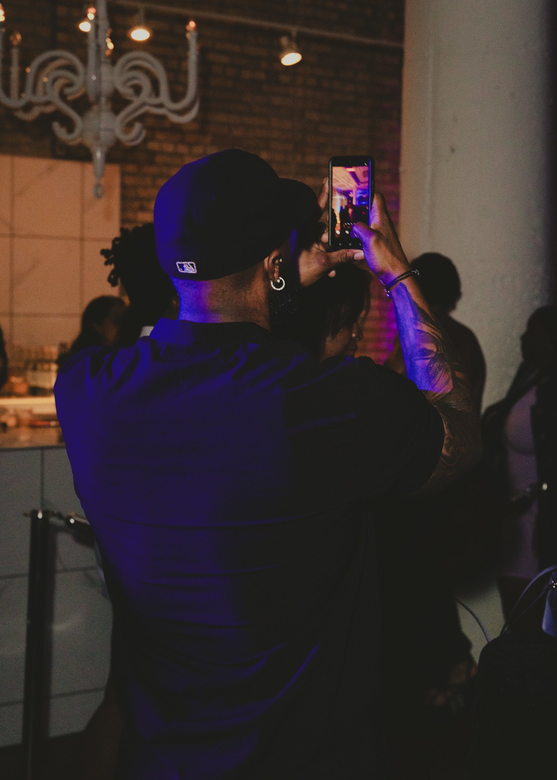 Person wearing a black cap taking a photo or video with a smartphone at an indoor event with dim lighting and a chandelier.
