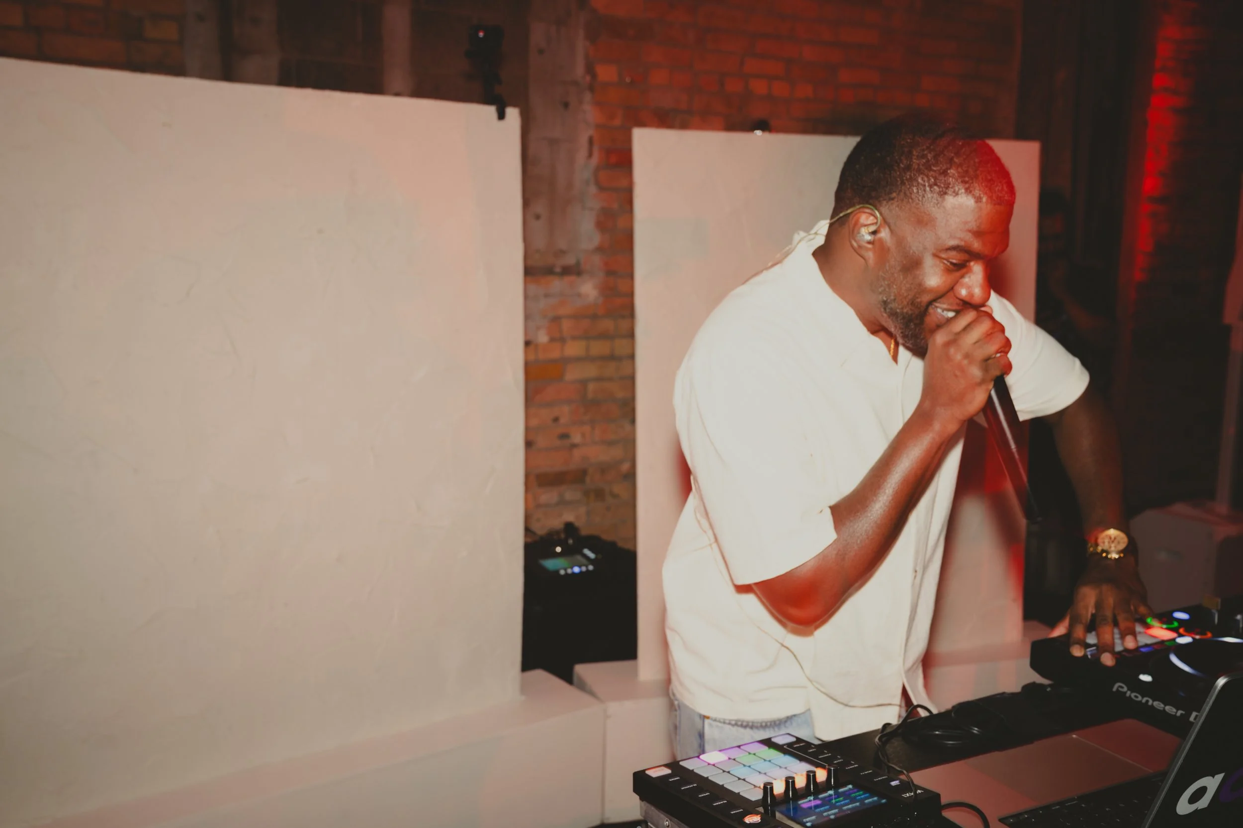A man smiling and laughing while holding a microphone, standing at a DJ mixing table with electronic music equipment, in a brick-walled venue with minimal decoration and colorful lighting.