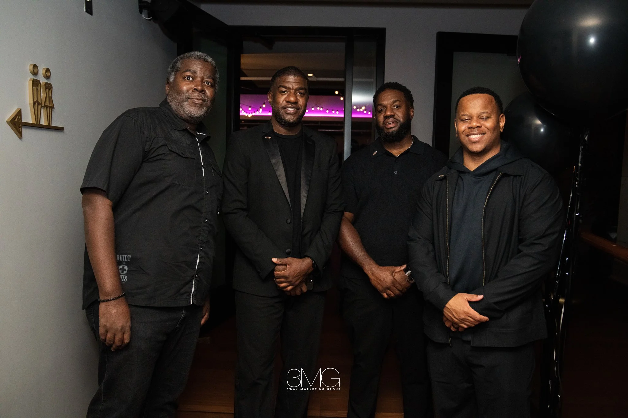 Four men standing indoors, dressed in dark clothing, posing for a photo. There are black balloons on the right side. The background shows a room with purple and pink lighting.