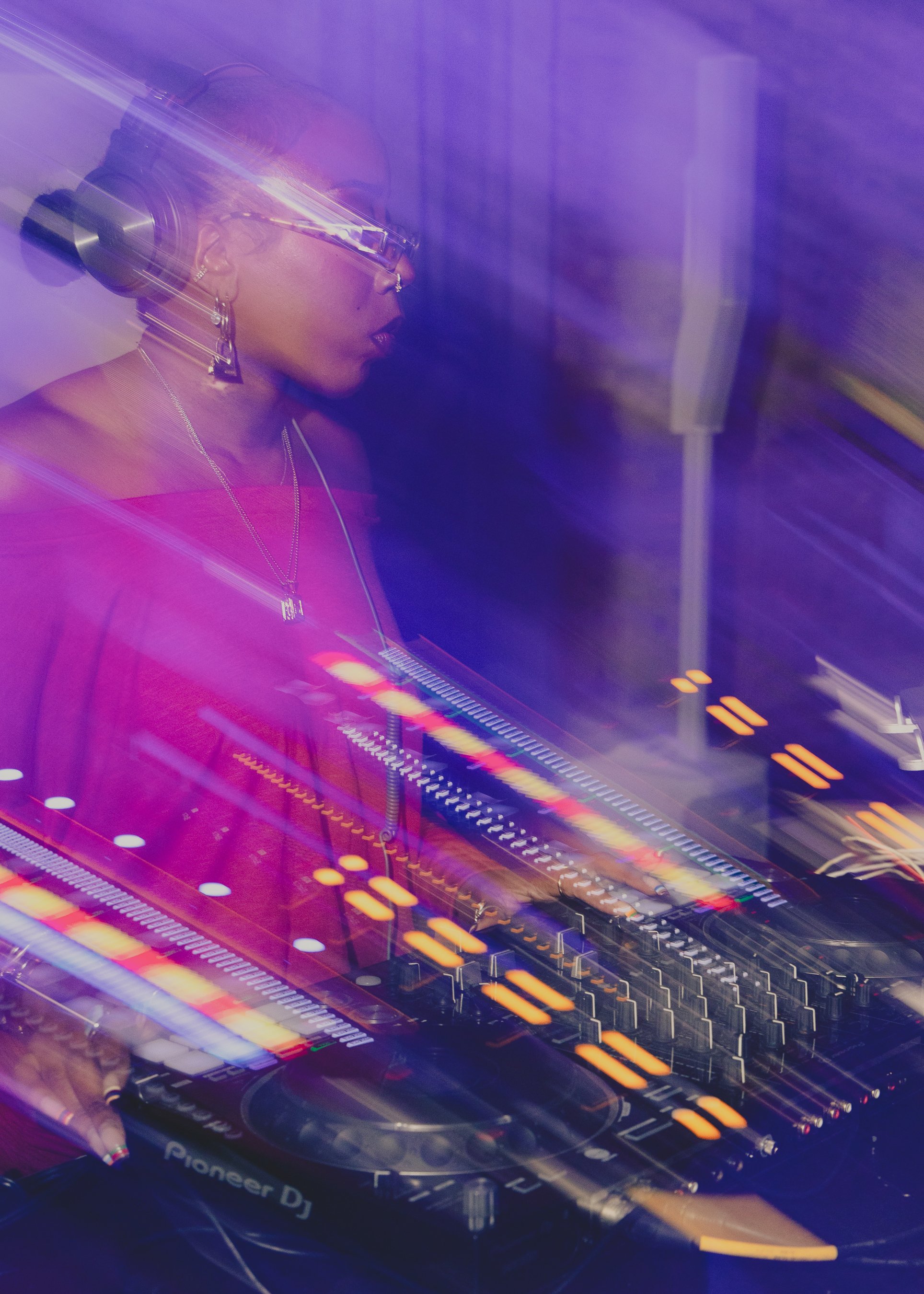 A female DJ wearing glasses and earrings mixes music on a Pioneer DJ controller in a dimly lit, purple-hued setting with motion blur effects.