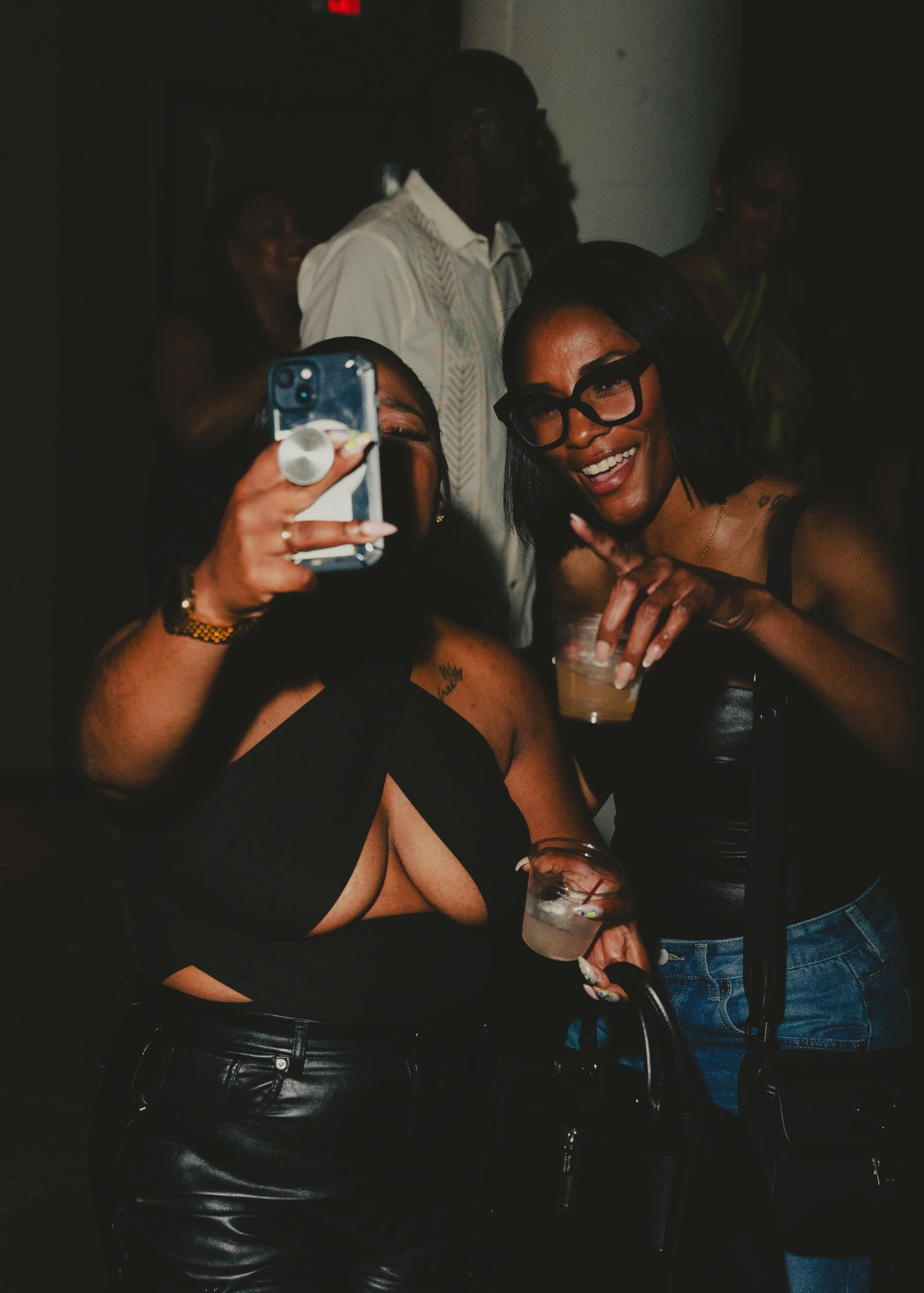 Two women taking a selfie at a party, one woman wearing glasses, holding a drink, and smiling while the other woman is holding a phone to capture the photo.