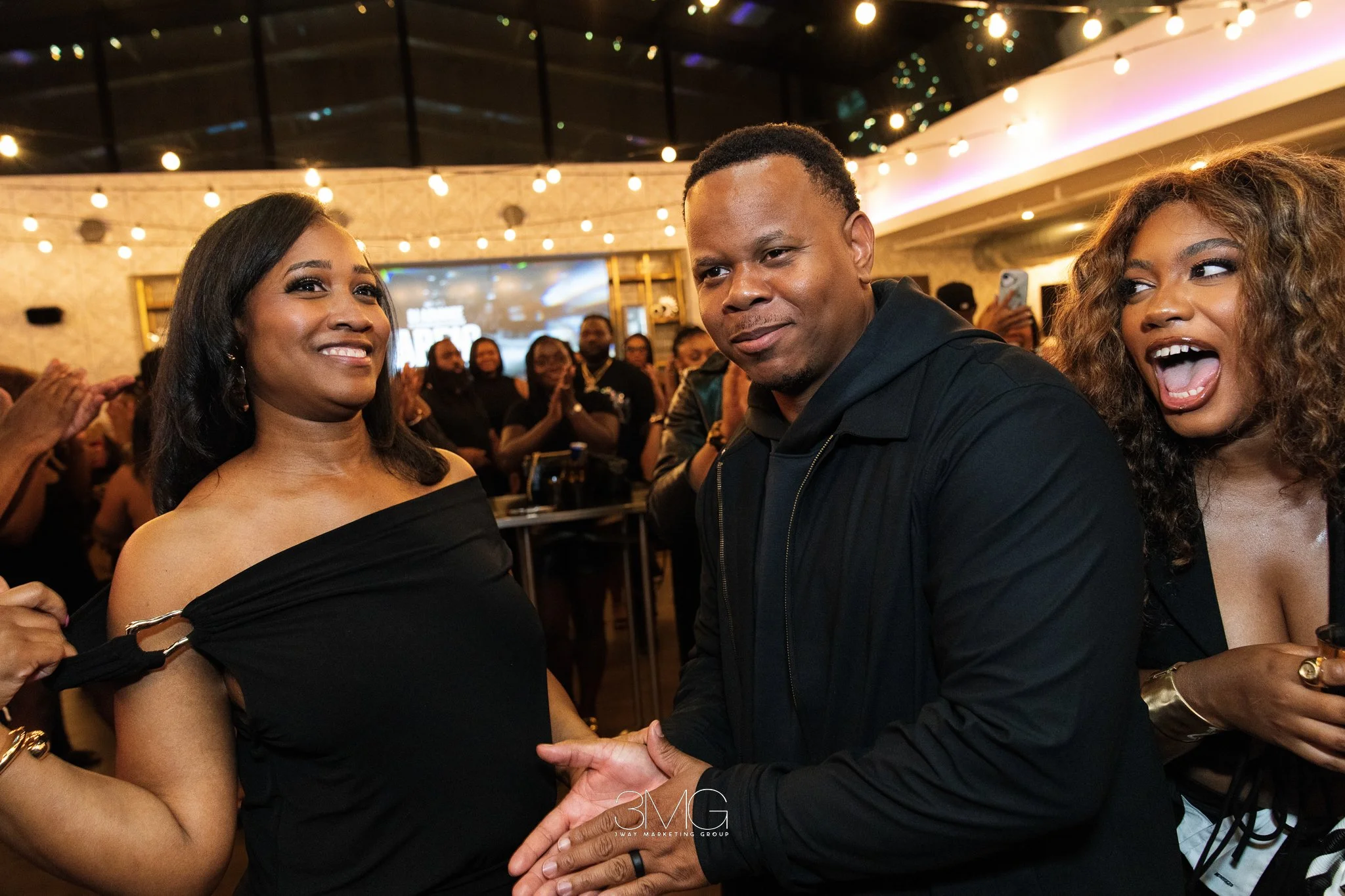 A group of people at an indoor event, with two women and one man in the foreground. The woman on the left has dark hair and is wearing a black off-the-shoulder dress and smiling. The man in the middle has short curly hair and is wearing a black jacke