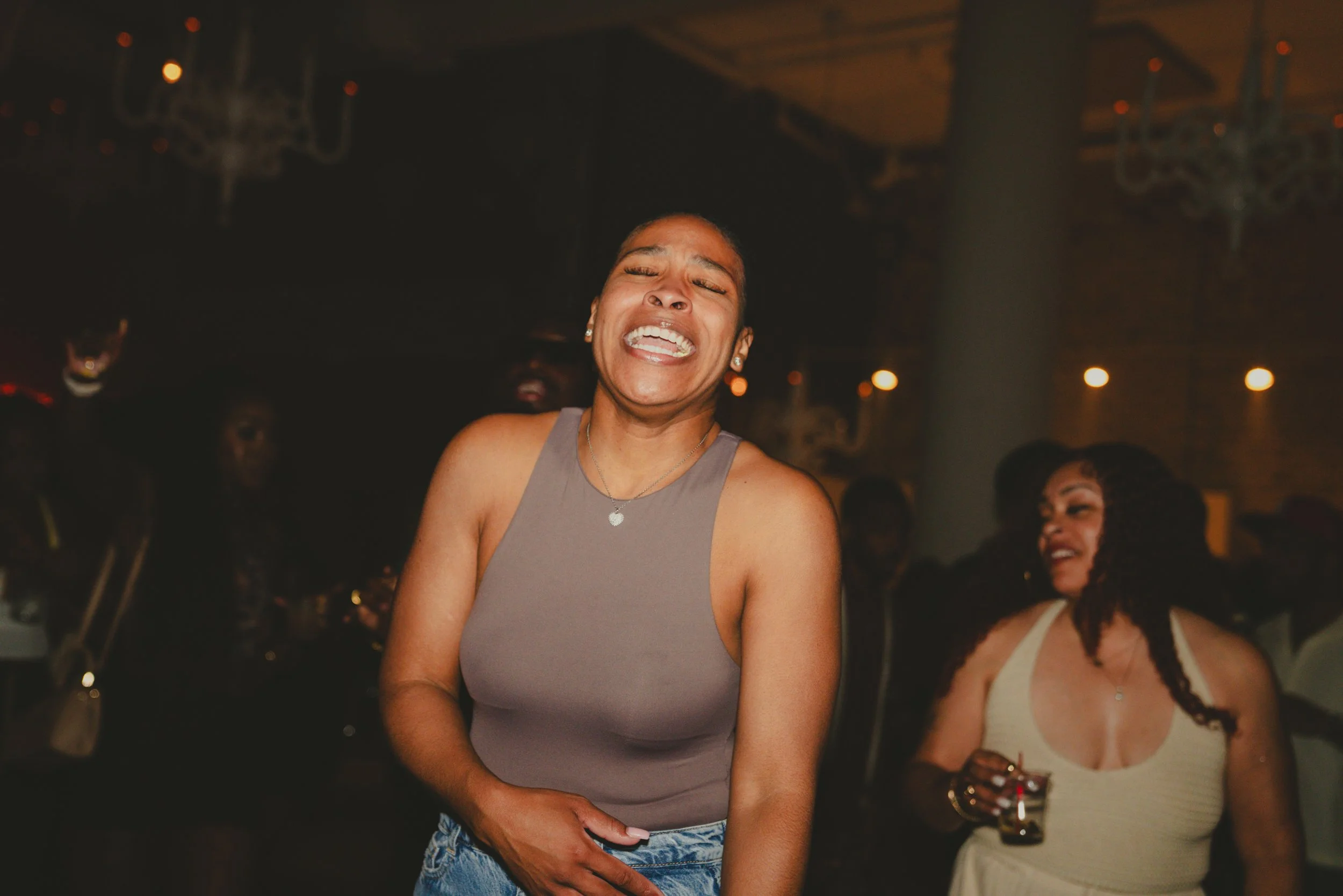 A woman with a big smile, closed eyes, and wearing a gray tank top, earrings, and a necklace, standing at a lively indoor party or event with other people in the background.