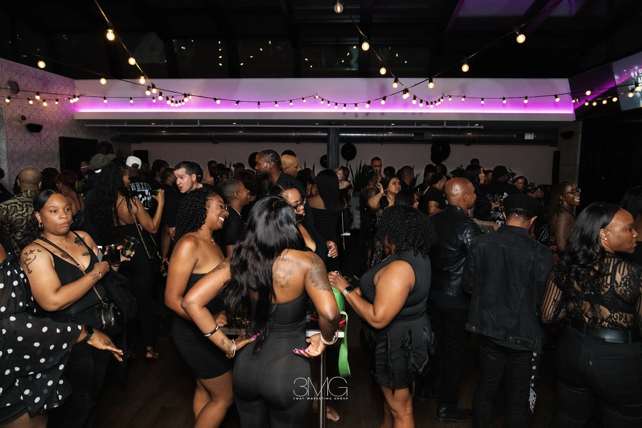 A large crowd of people gathered at an indoor event or party, with several women and men dressed in black, some holding drinks, under string lights and purple lighting.