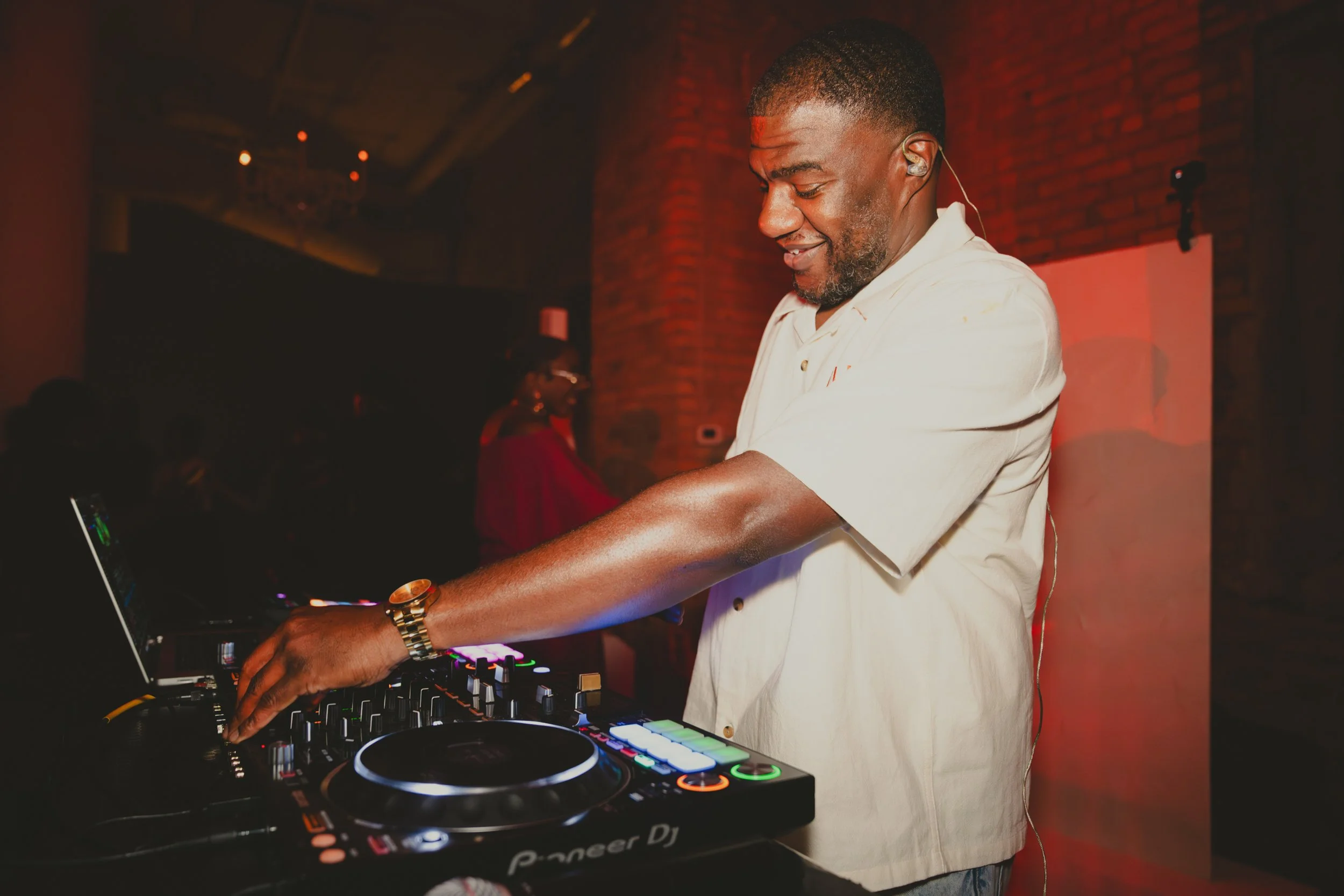 A man is DJing at a party, wearing a beige shirt and a gold watch, with a focused expression on his face as he operates a DJ controller.