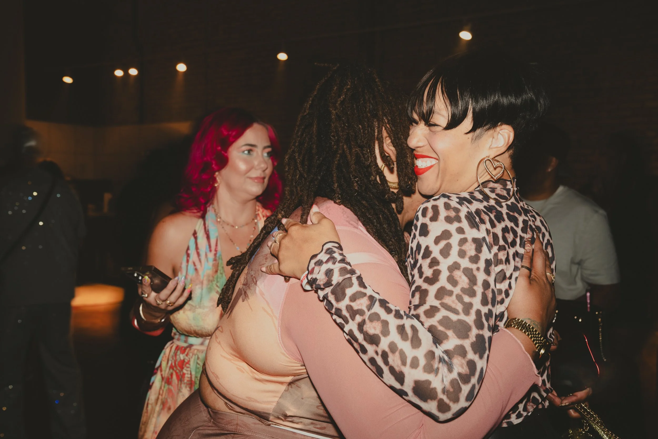 Two women hugging and smiling at each other at a social event, with other people in the background.