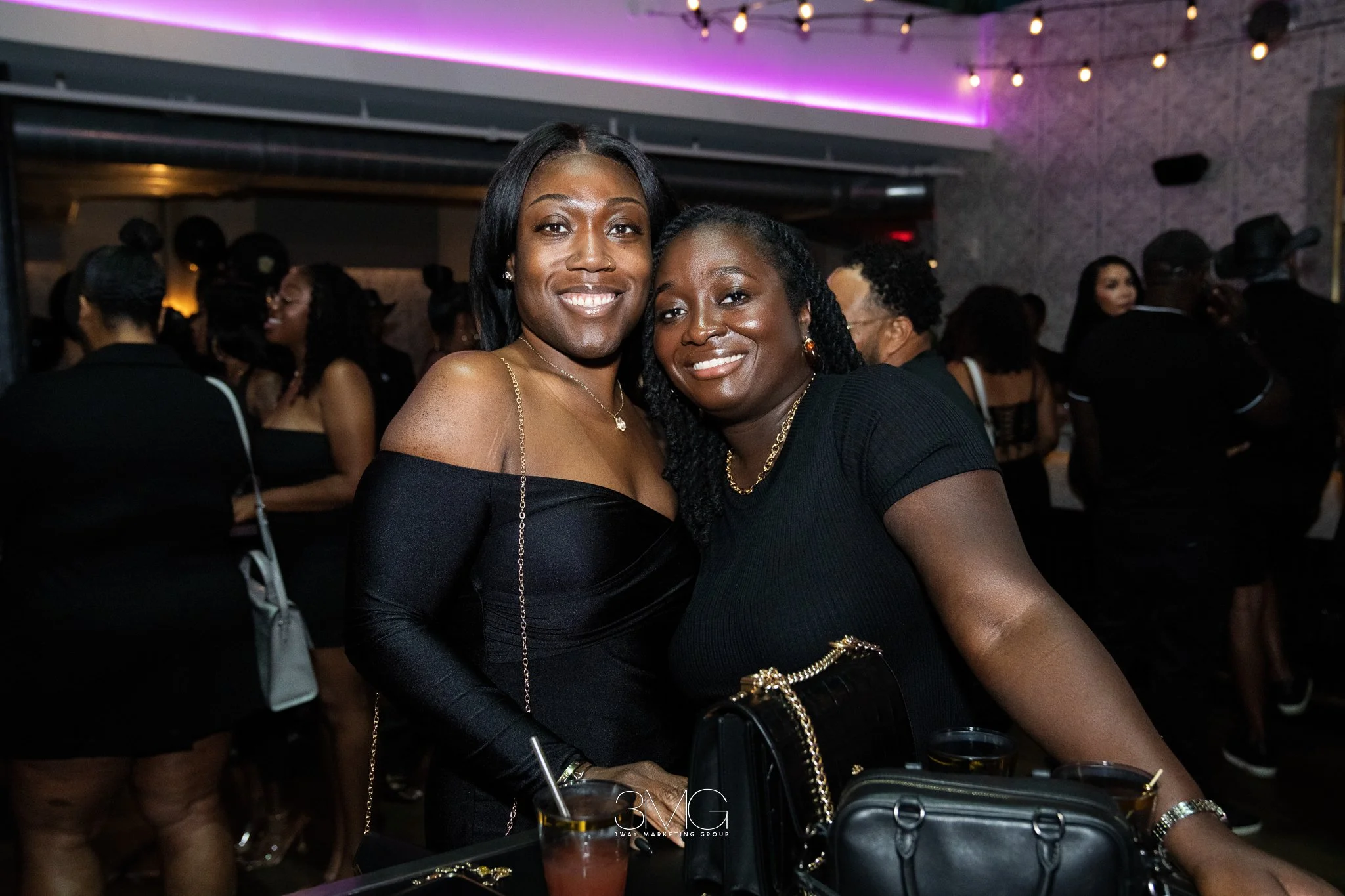 Two women smiling and posing for a photo at a lively party or social gathering. They are dressed elegantly, with one wearing an off-shoulder black dress and the other in a black top. The background shows other guests socializing, with some blurred ou