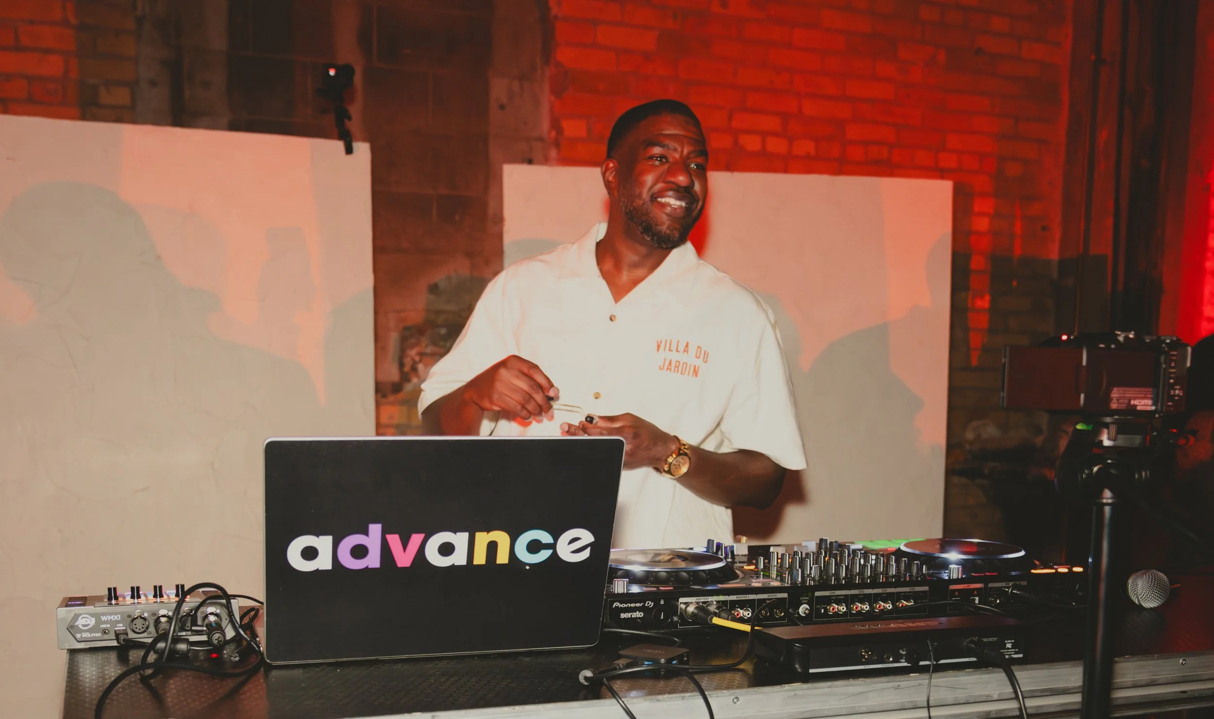A man with a beard and short hair, wearing a white shirt with 'VILLA DU JARDIN' written on it, stands behind DJ equipment at a party or event, smiling slightly. There is a laptop with an 'advance' sticker, DJ controllers, and a camera on a tripod nea