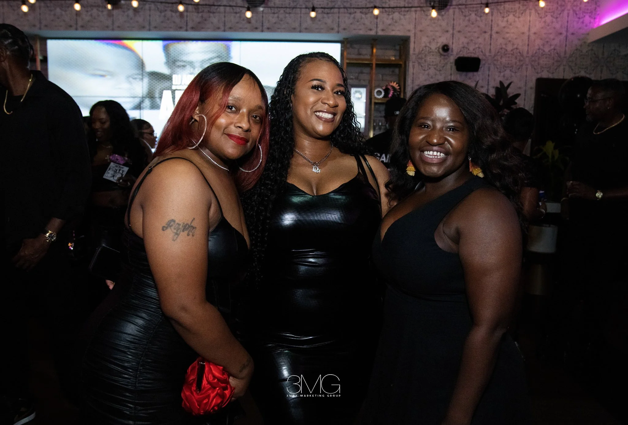Three women smiling, standing close together at a social event or party, with a brightly lit background and people in the background.